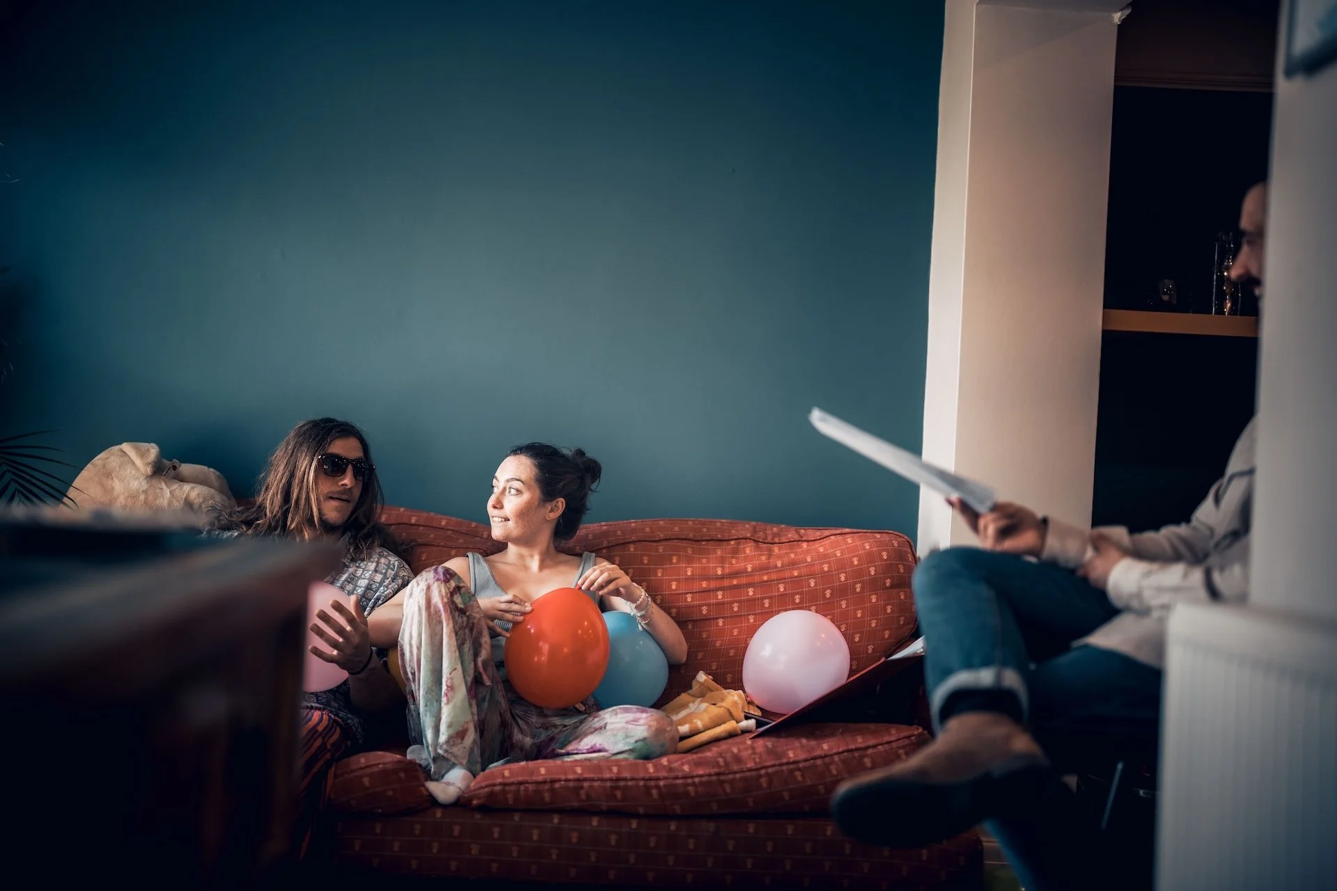 Three people are sitting on a red couch with balloons and snacks around them, during a casual get-together. One person is wearing sunglasses and a patterned shirt, another a tank top, and the third person is holding papers while sitting nearby.