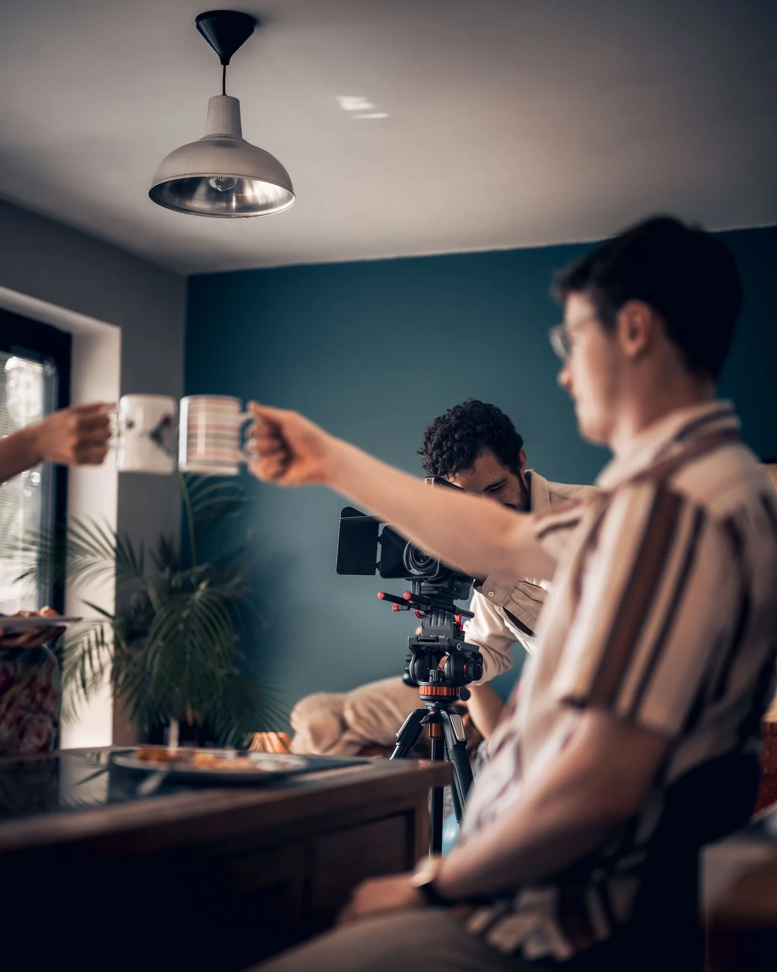 Two men working with a video camera on a tripod in a room with teal walls and a ceiling light, one person holding a mug, plants and window in the background.