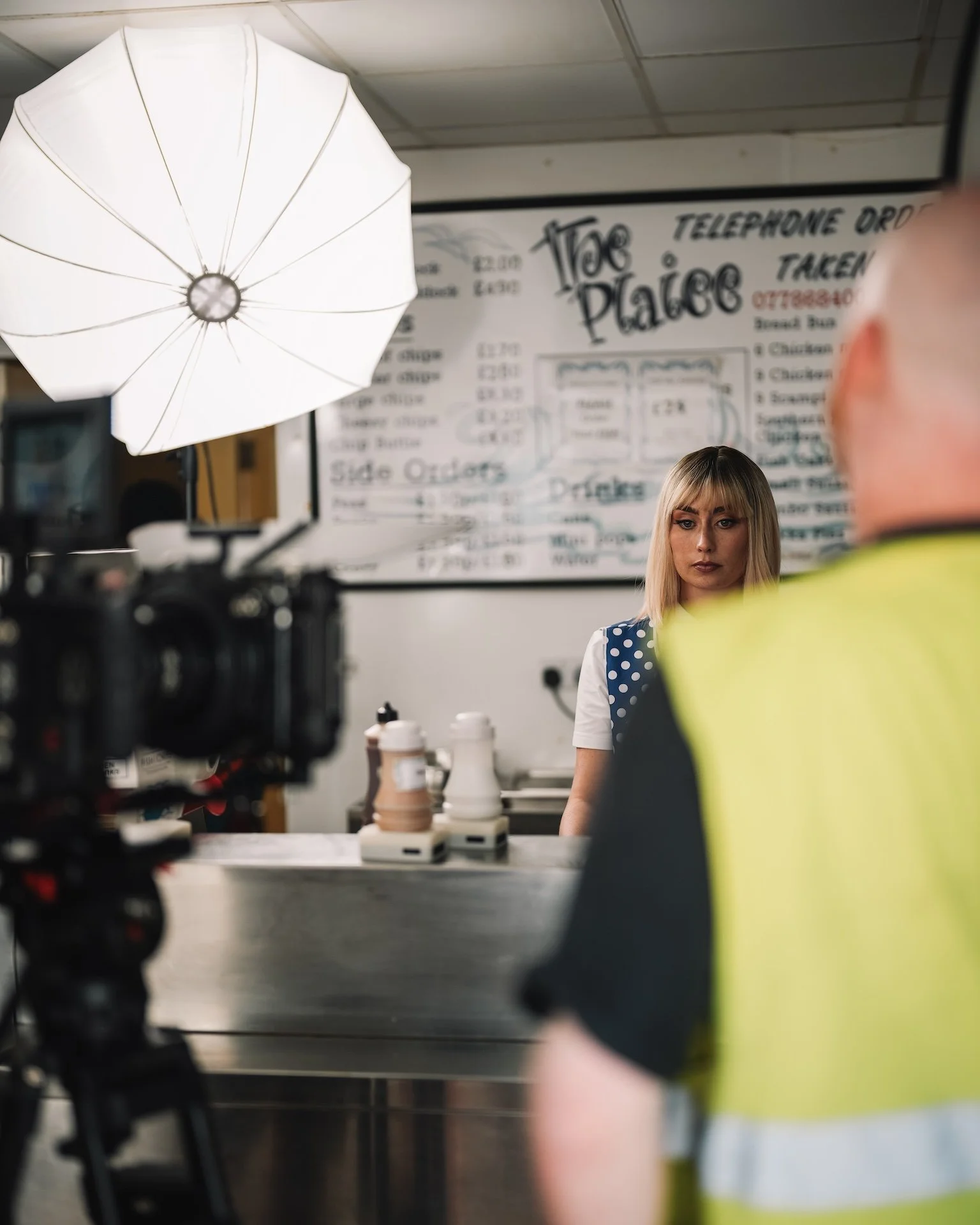 A woman with blonde hair and a polka dot dress stands behind a counter in a restaurant, with a whiteboard menu visible in the background. There is a large photography umbrella and camera equipment in the foreground.