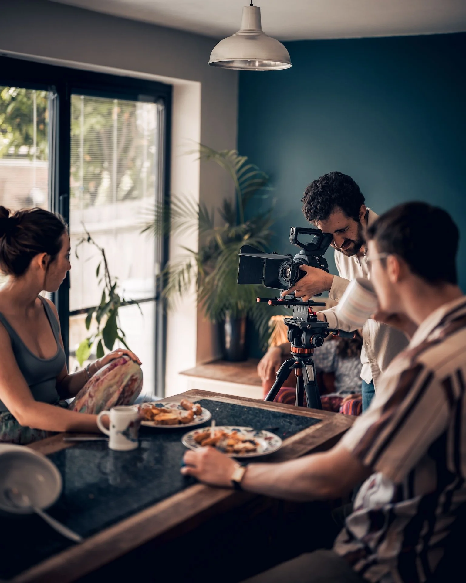 People filming a scene in a kitchen, with a woman holding a colorful cloth, a man checking a camera, and others at the table with pizza and coffee.