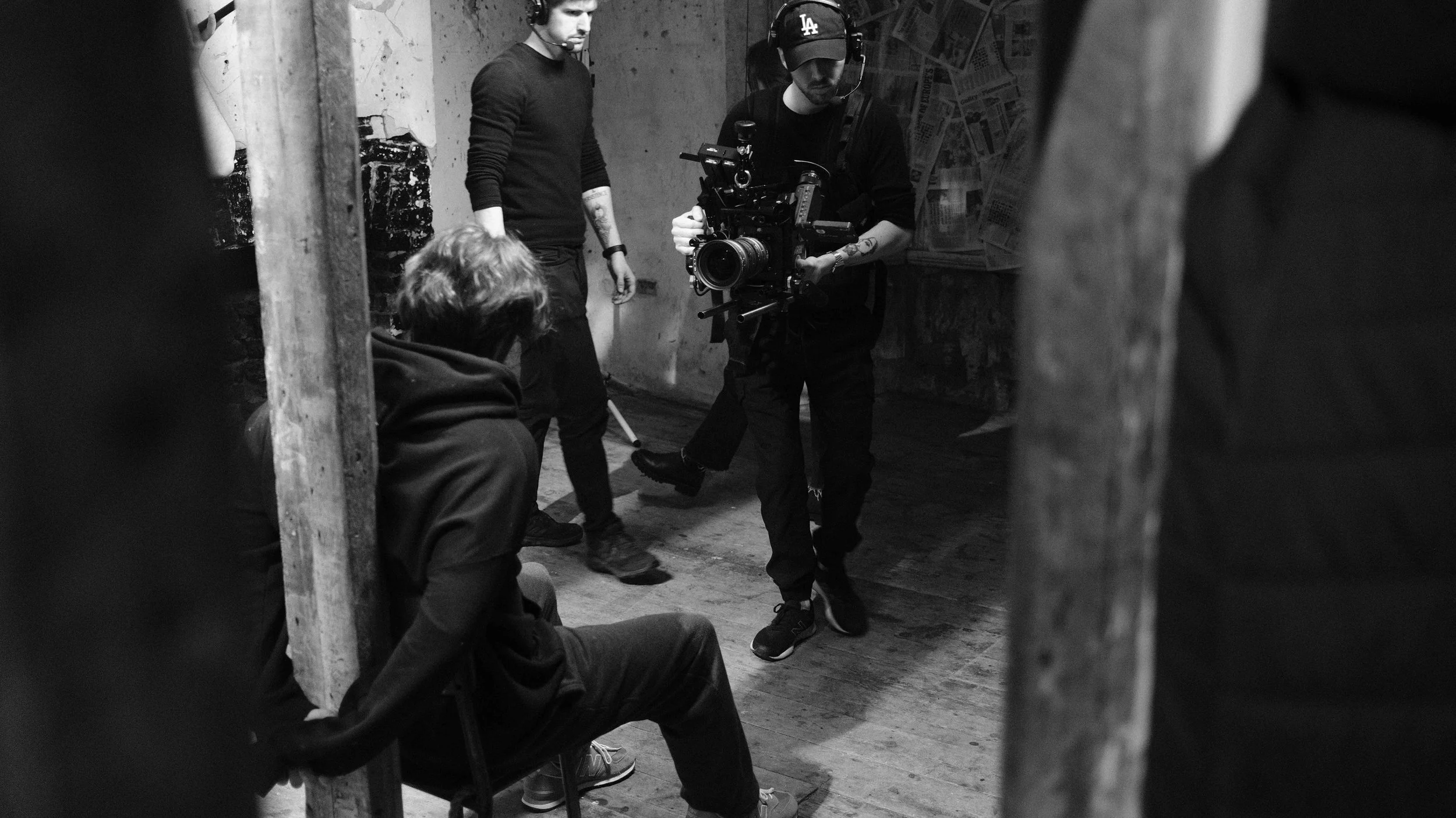 Black and white image of a film crew on a set, with one person sitting and another standing, while a cameraman films them. The scene is indoors with a worn wall and newspapers or posters on the background.