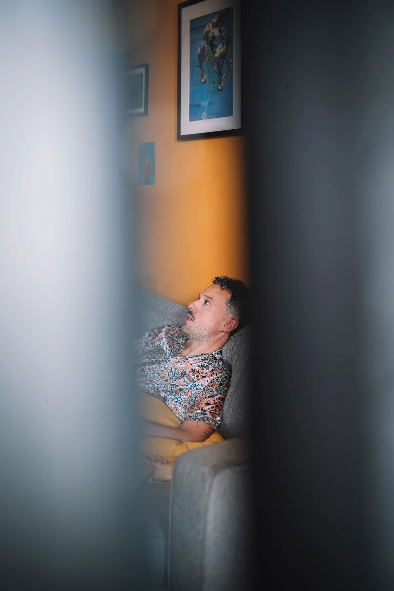 A man sitting on a gray couch, looking up, with framed artwork on the wall behind him, and the photo is taken through a partly blurred, overlapping frame.