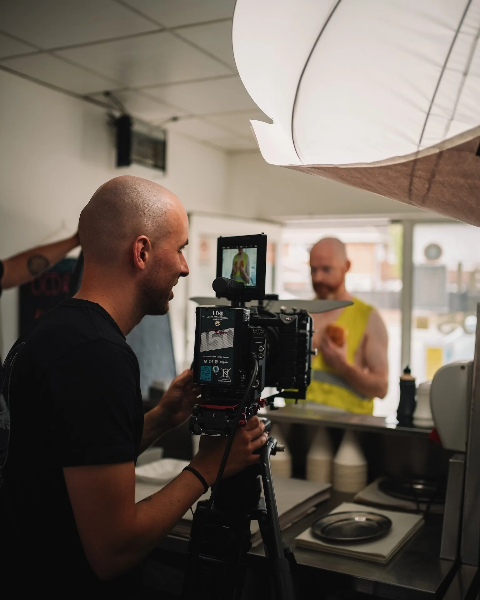 A man operating a camera on a tripod in a kitchen, filming a woman in a yellow top who is holding a phone.