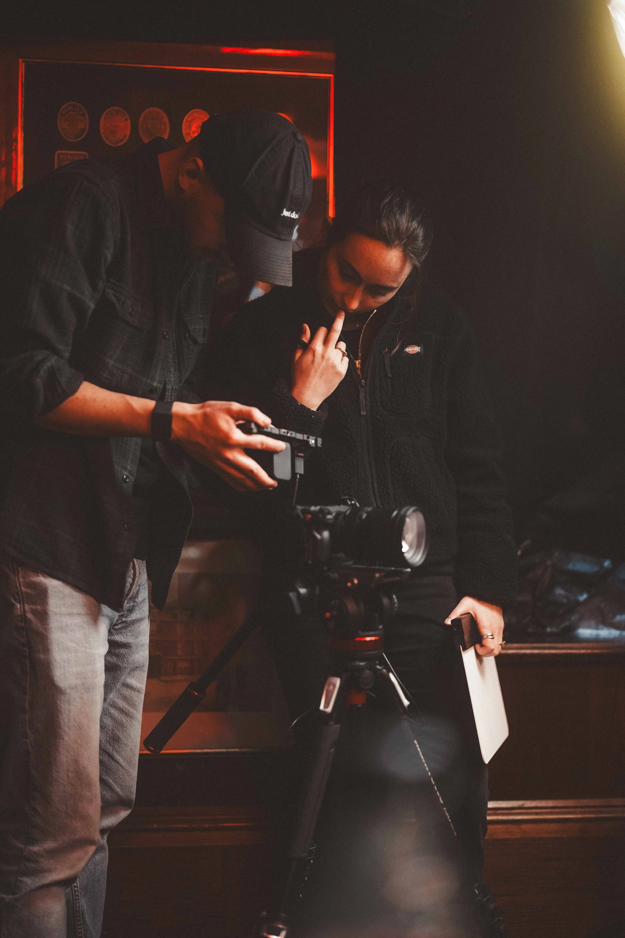 Two people are discussing something in a dimly lit room with a dark background. One person is wearing a checkered shirt and cap, holding a camera, and looking at something the other person is holding or pointing to. The second person has dark hair, i