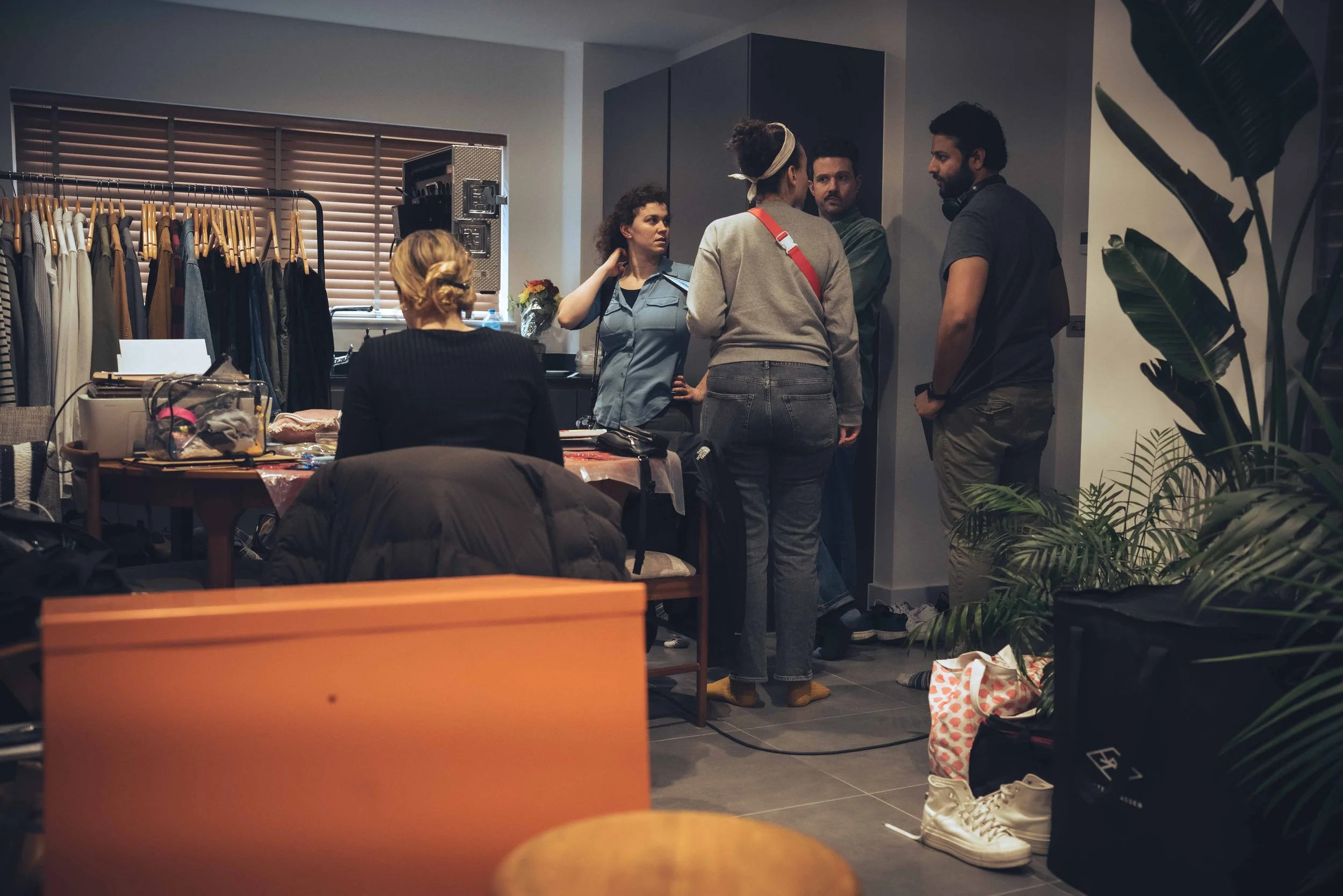 People gathered in a room, possibly a dressing room or backstage area, with clothing racks, a table with items, and plants in the foreground.
