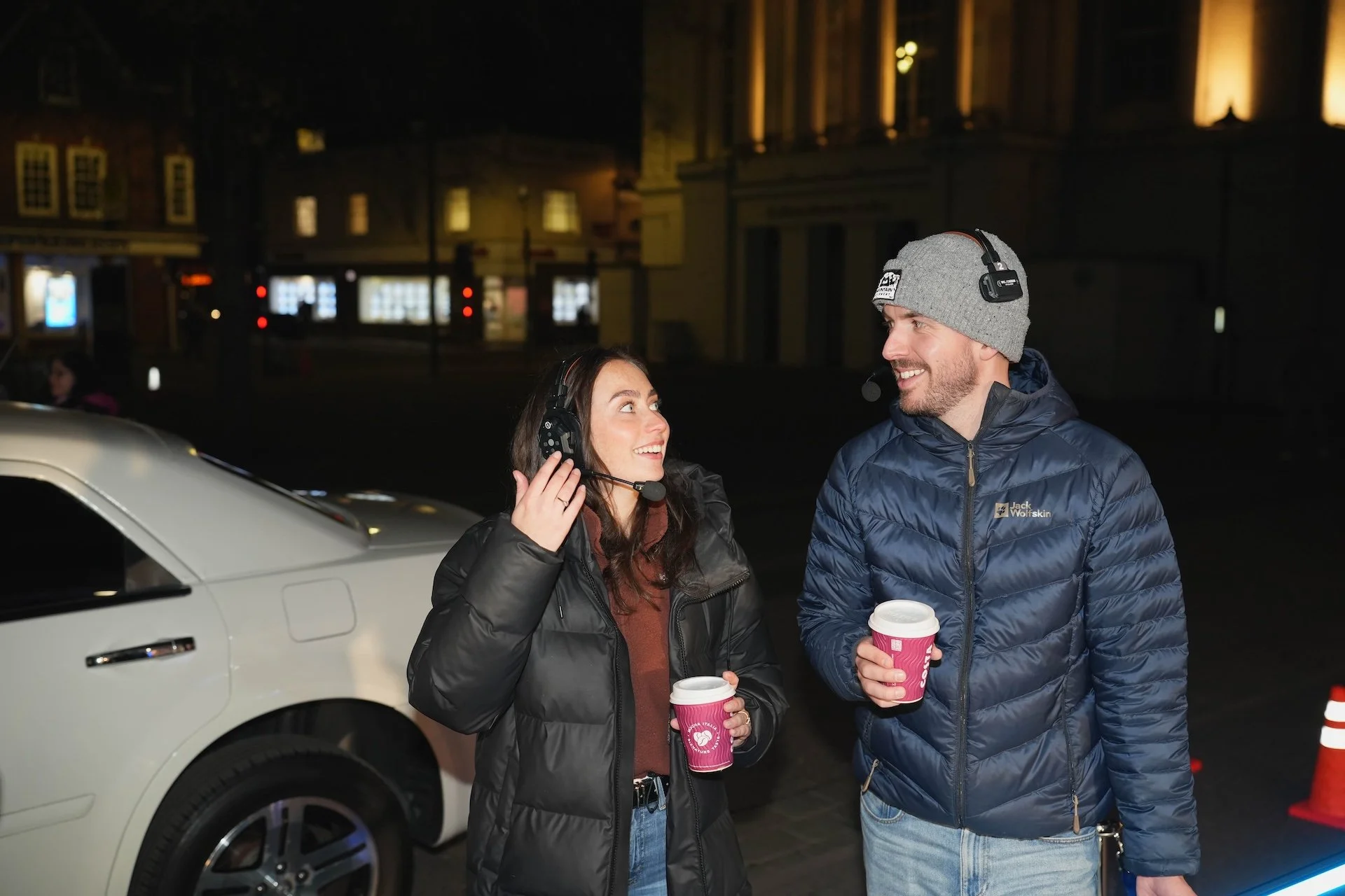 Two people stand outdoors at night, smiling and talking while holding coffee cups. They are wearing jackets and headphones, with a white car and illuminated buildings in the background.