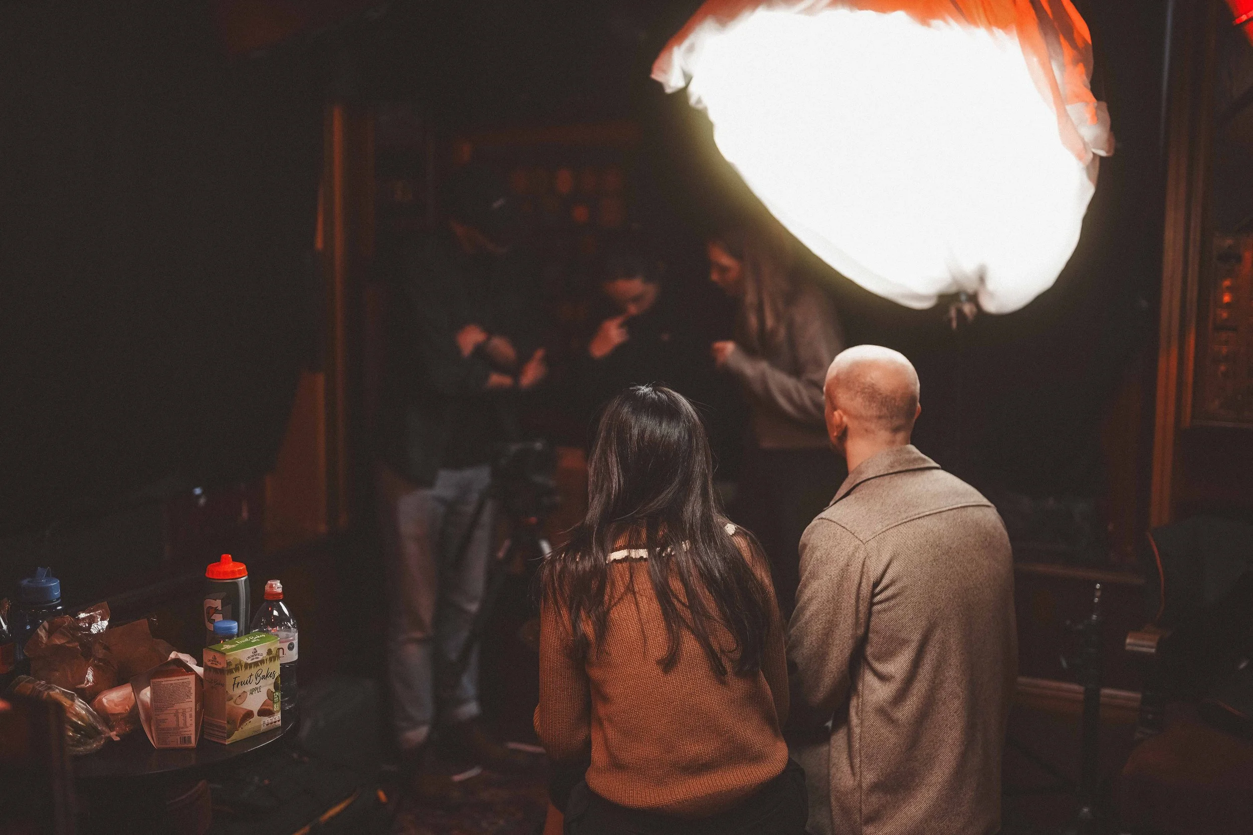 People gathered at a table in a dimly lit room, with some food and drinks on the table, and a large bright light or projection screen behind them, indicating a social event or gathering.