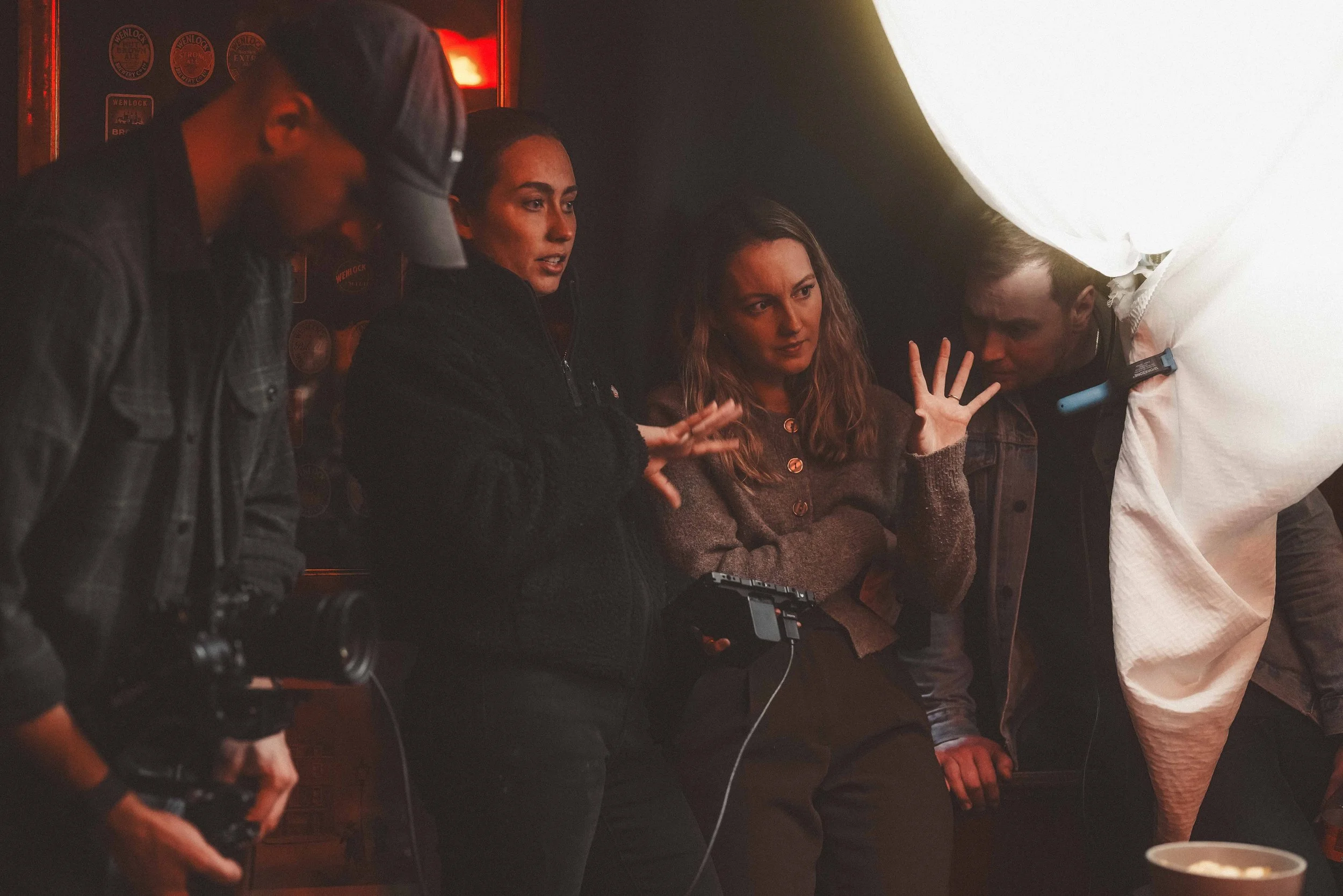 Four people gathered around a camera, discussing something. They are in a low-light environment with a large light source illuminating part of the scene.