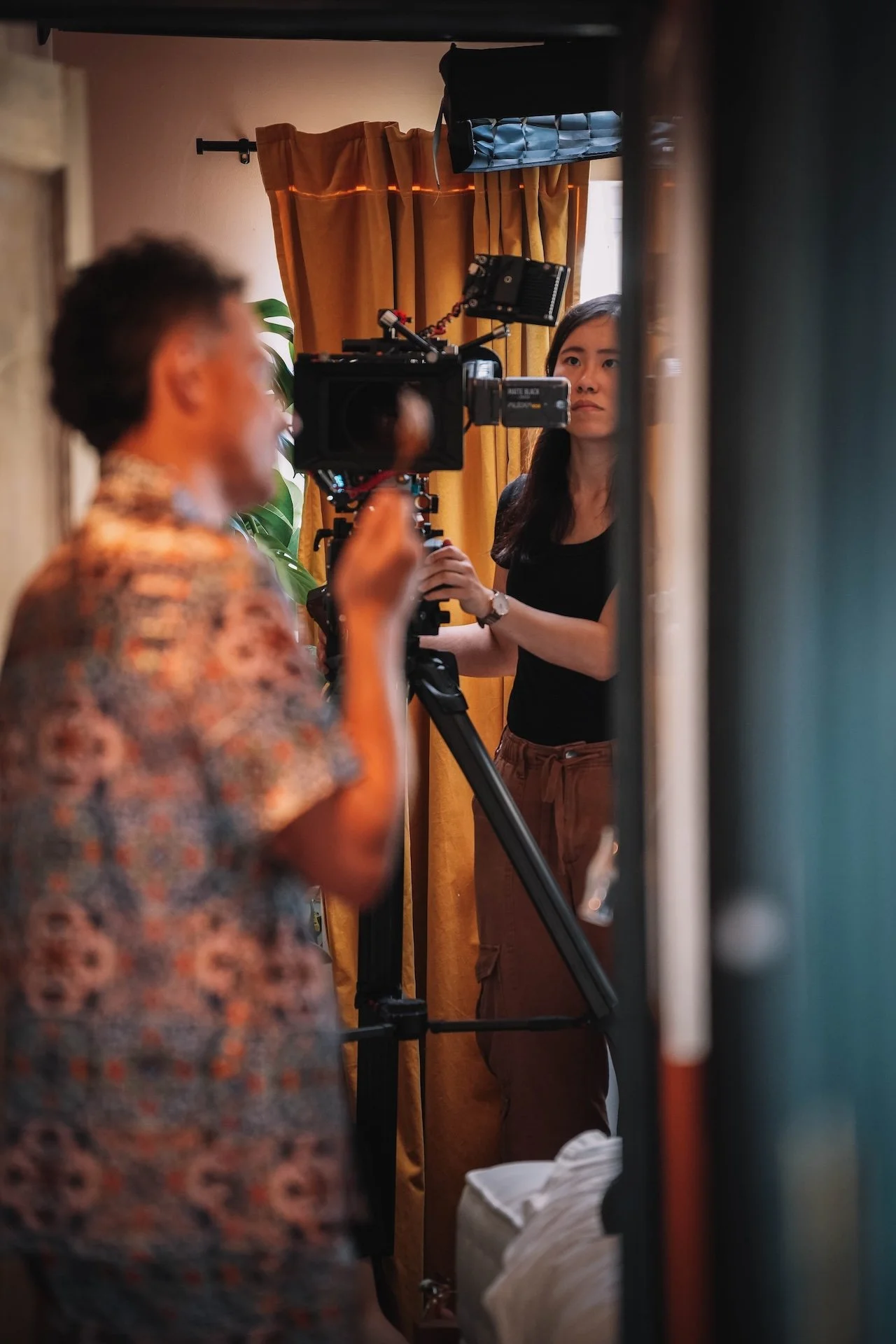 A woman is operating a camera on a tripod, capturing a man in a patterned shirt during what appears to be a filming or photography session in a room with brown curtains and a plant.
