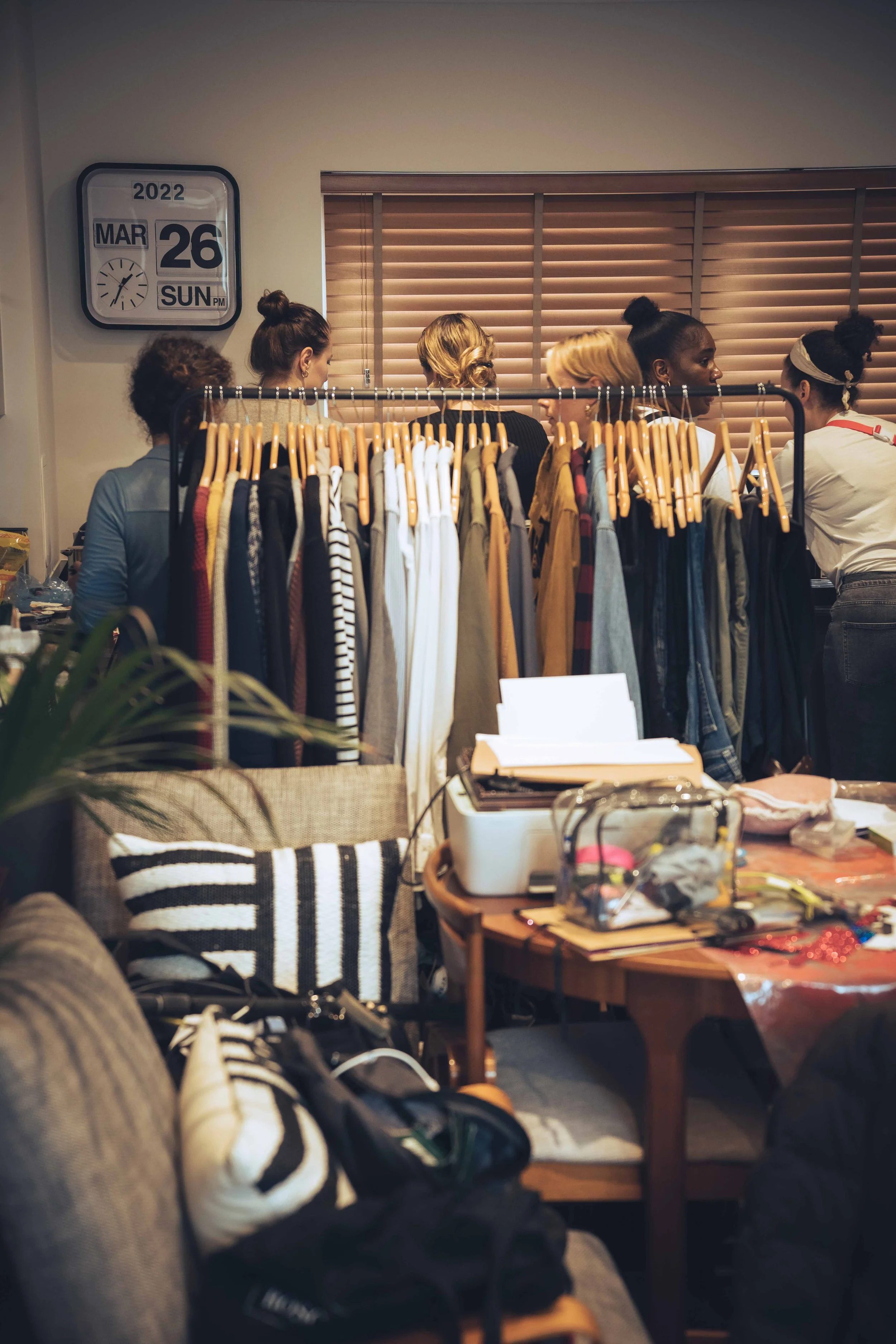A group of women standing behind a clothing rack filled with various shirts and dresses, engaging in conversation in a room with wooden blinds and a wall clock showing the date March 26, 2022.