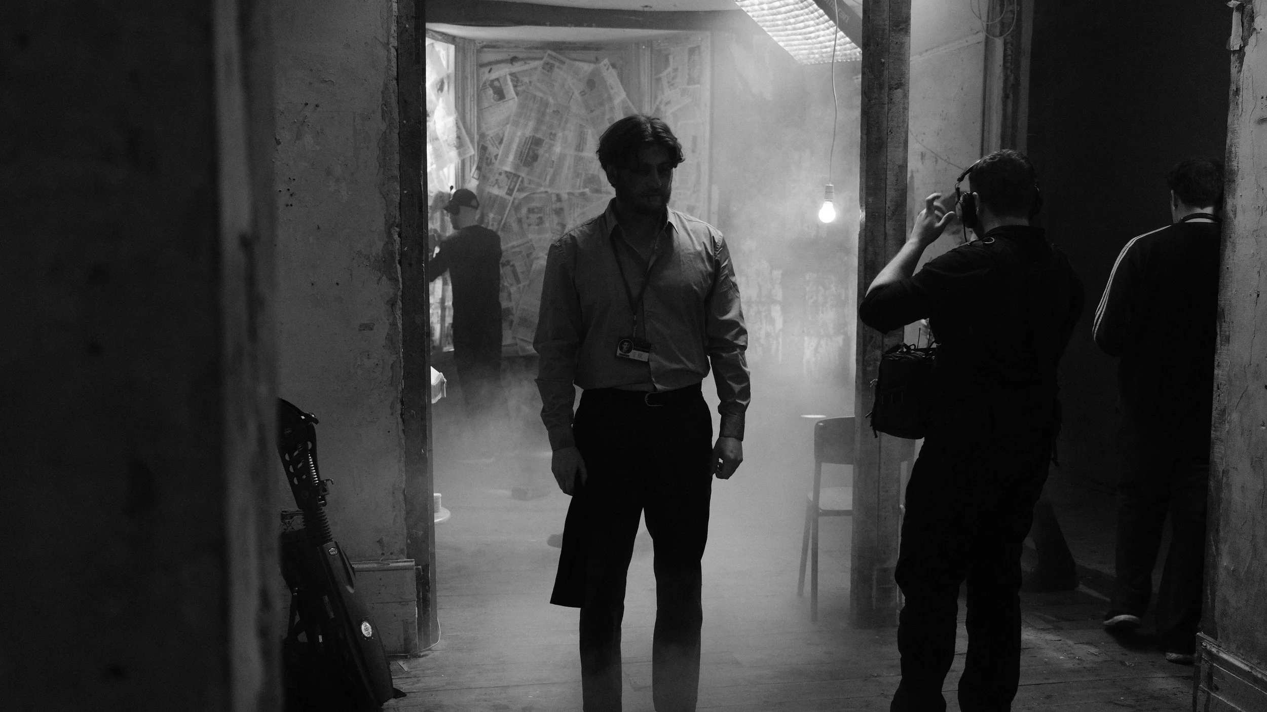 Black and white photo of a man with a badge standing in a smoky room, while a second person wearing headphones takes a photo or video of him. Several other people are in the background, with a wall covered in newspapers.