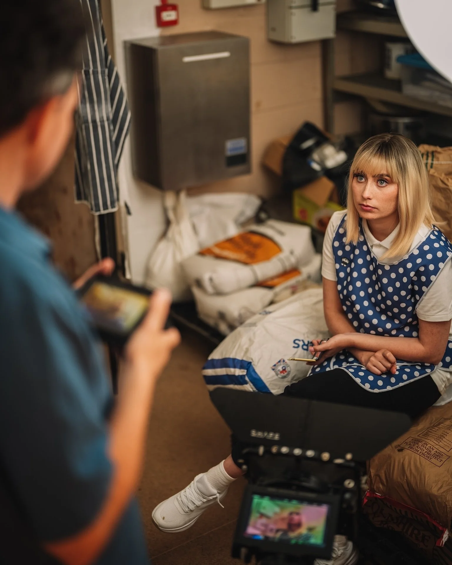 A woman sits on the floor in a room, wearing a white shirt and a polka dot vest, being photographed or filmed by a person holding a camera and a phone, with some equipment and supplies around them.