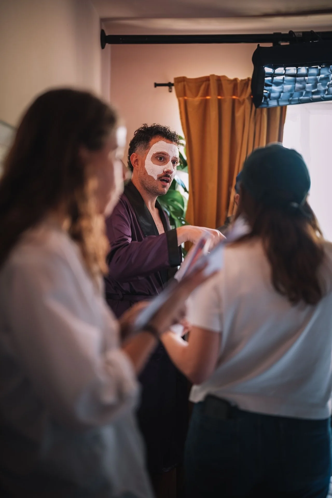 A man with face paint in white circles around his eyes and on his forehead, talking to two women in a room with curtains and lighting equipment visible.
