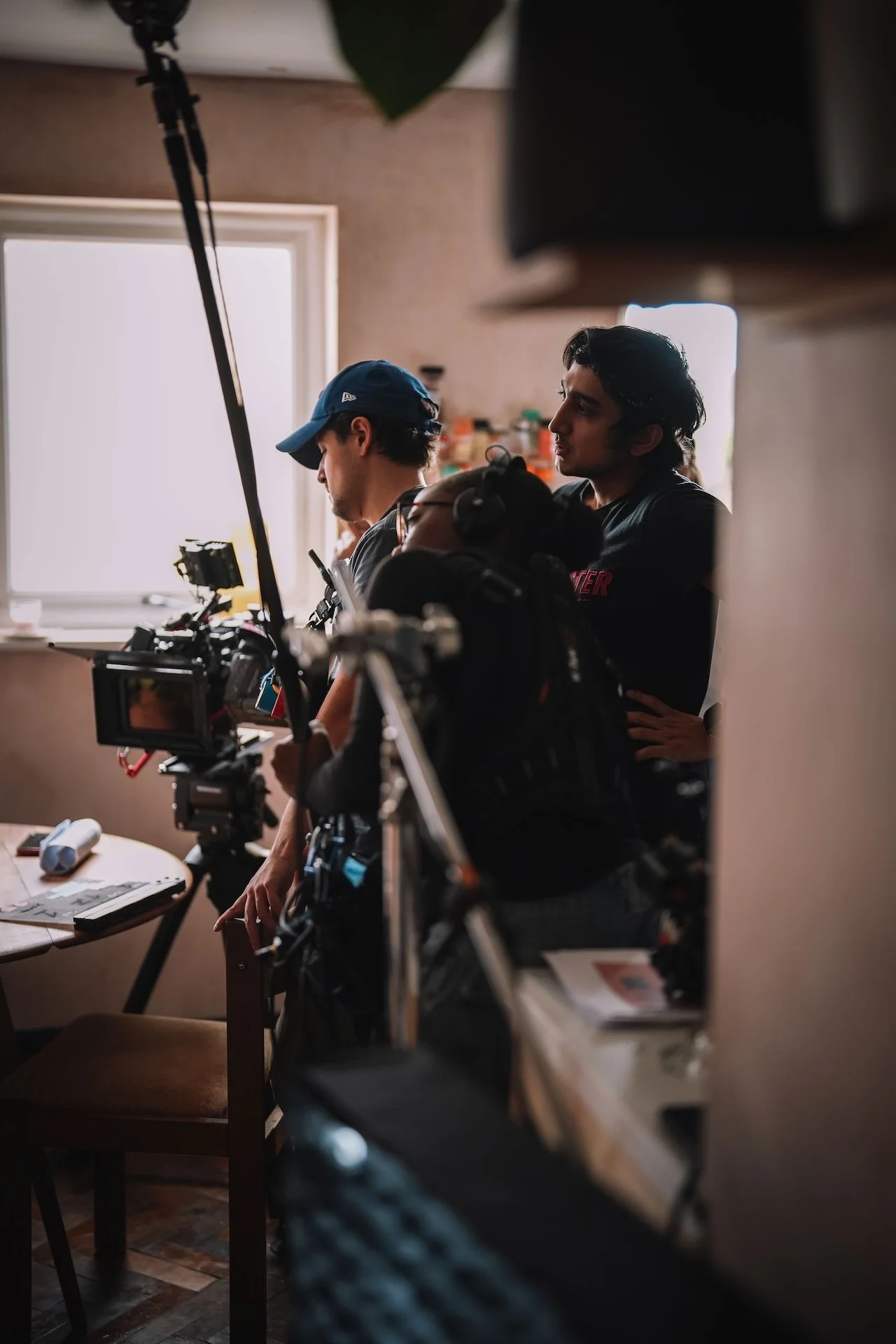 Behind-the-scenes shot of two men working in a room, viewed through a mirror or a frame, with a camera setup on a table in the foreground.