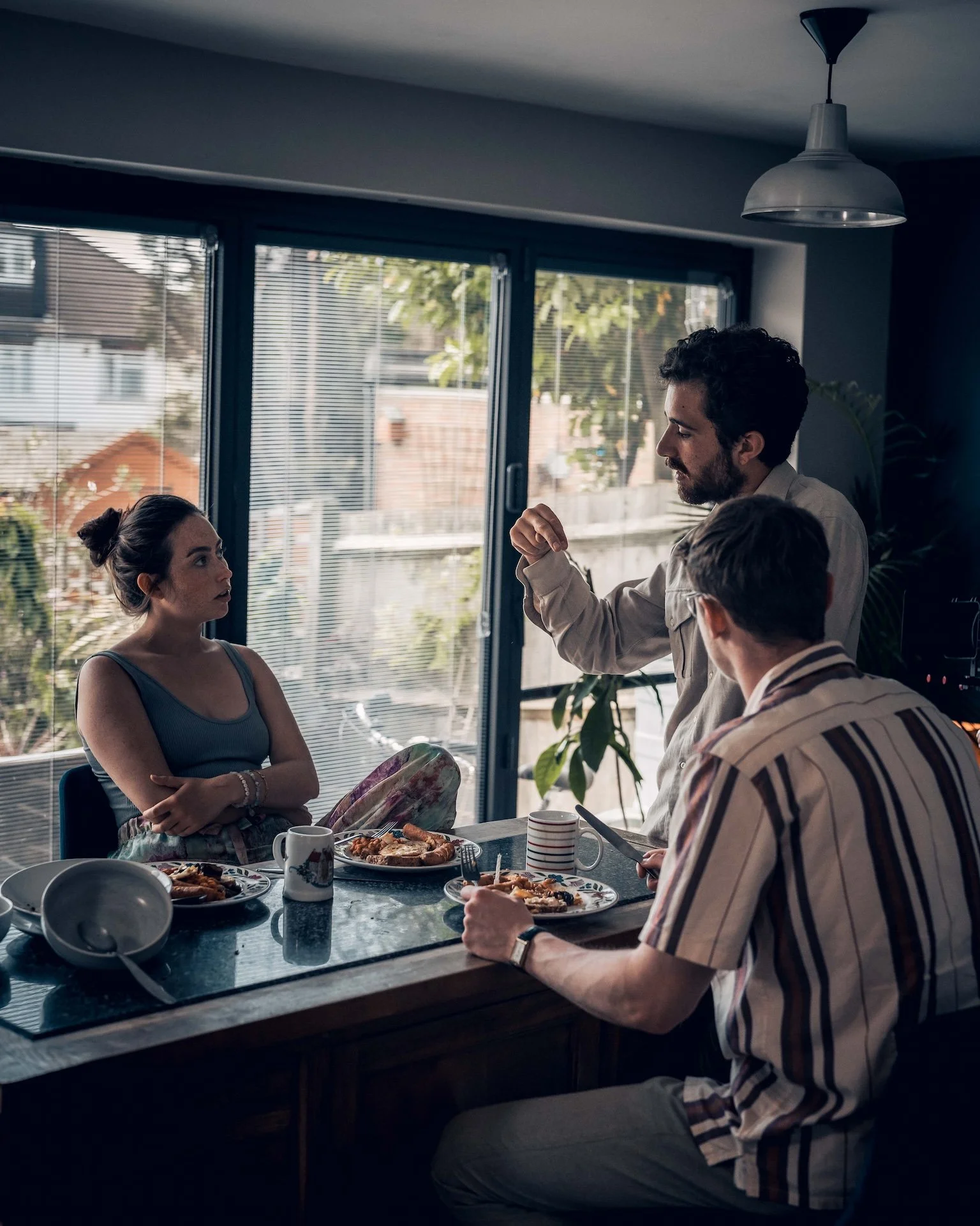 A group of four people having a meal at a kitchen table, with food, cups, and dishes, inside a room with large windows and blinds, with a man talking.