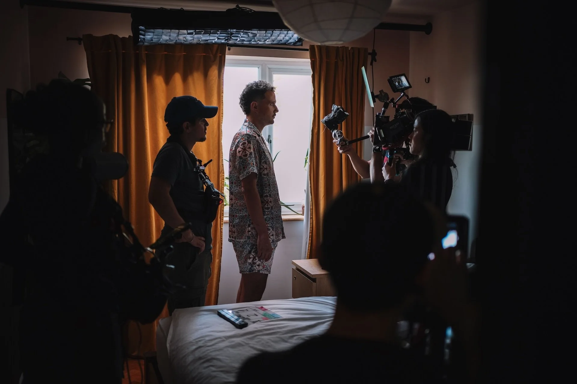 A film crew filming a person standing in a room with yellow curtains and a window, with equipment and crew members visible around.