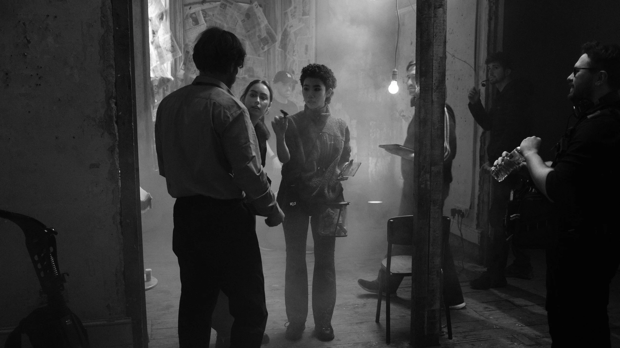 Behind-the-scenes of a film crew in a smoke-filled room with several crew members, including one woman holding a clapperboard and taking notes, and a man holding a water bottle, all engaged in a discussion.