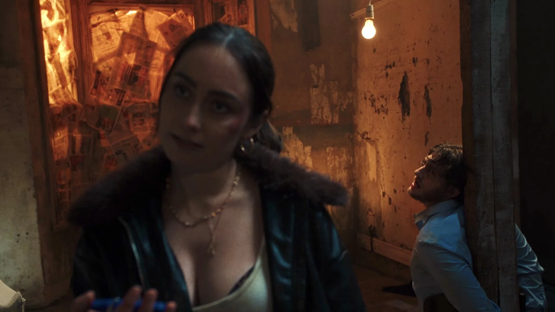 A woman with dark hair, earrings, and a pearl necklace stands in the foreground, looking to the side with a neutral expression, in a dimly lit room with a rough, worn wall and a man sitting on the floor in the background, leaning against a wooden beam, with a lit light bulb overhead.