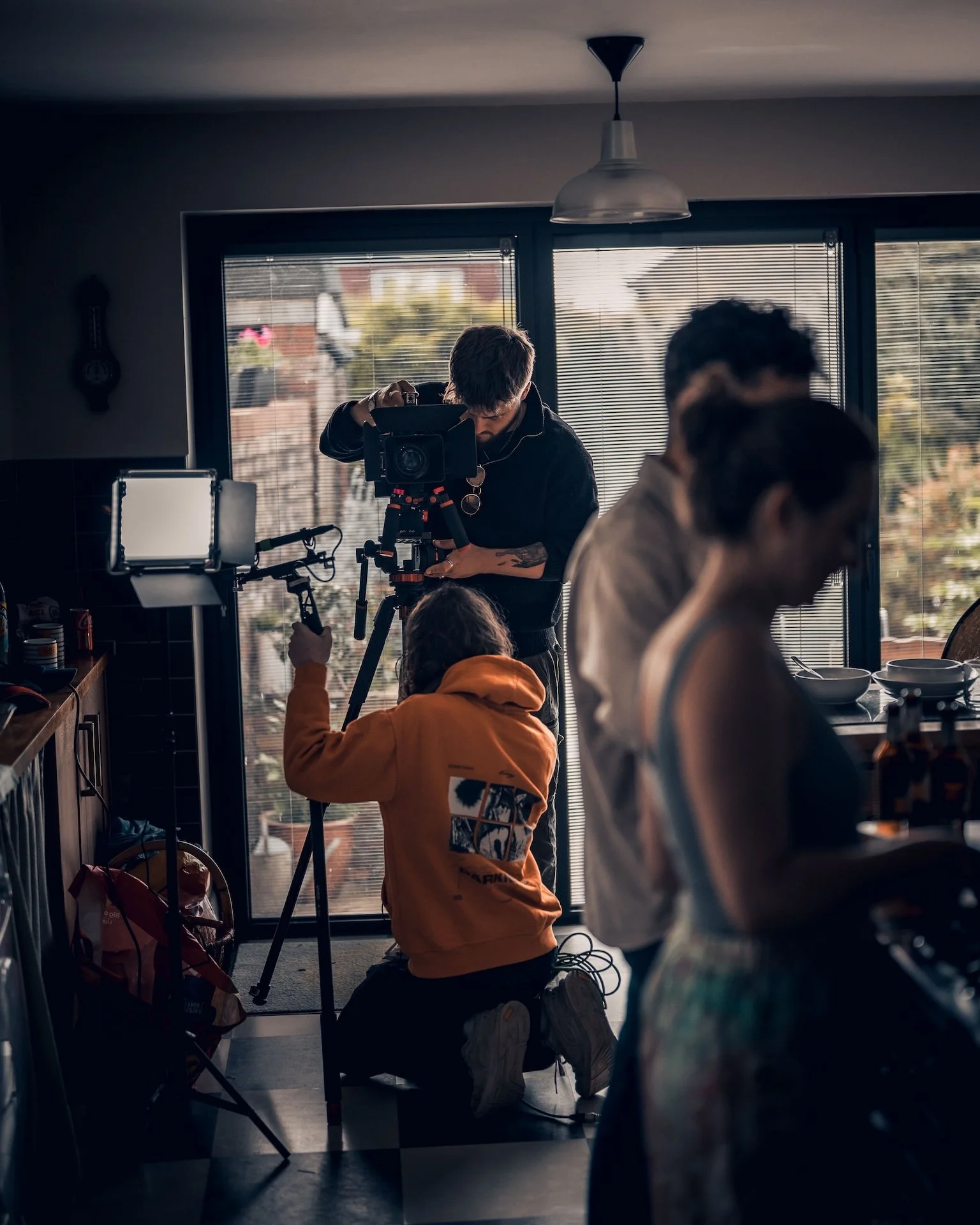 People filming in a cozy kitchen with large windows, a hanging light, and blinds, with outdoor greenery visible outside.