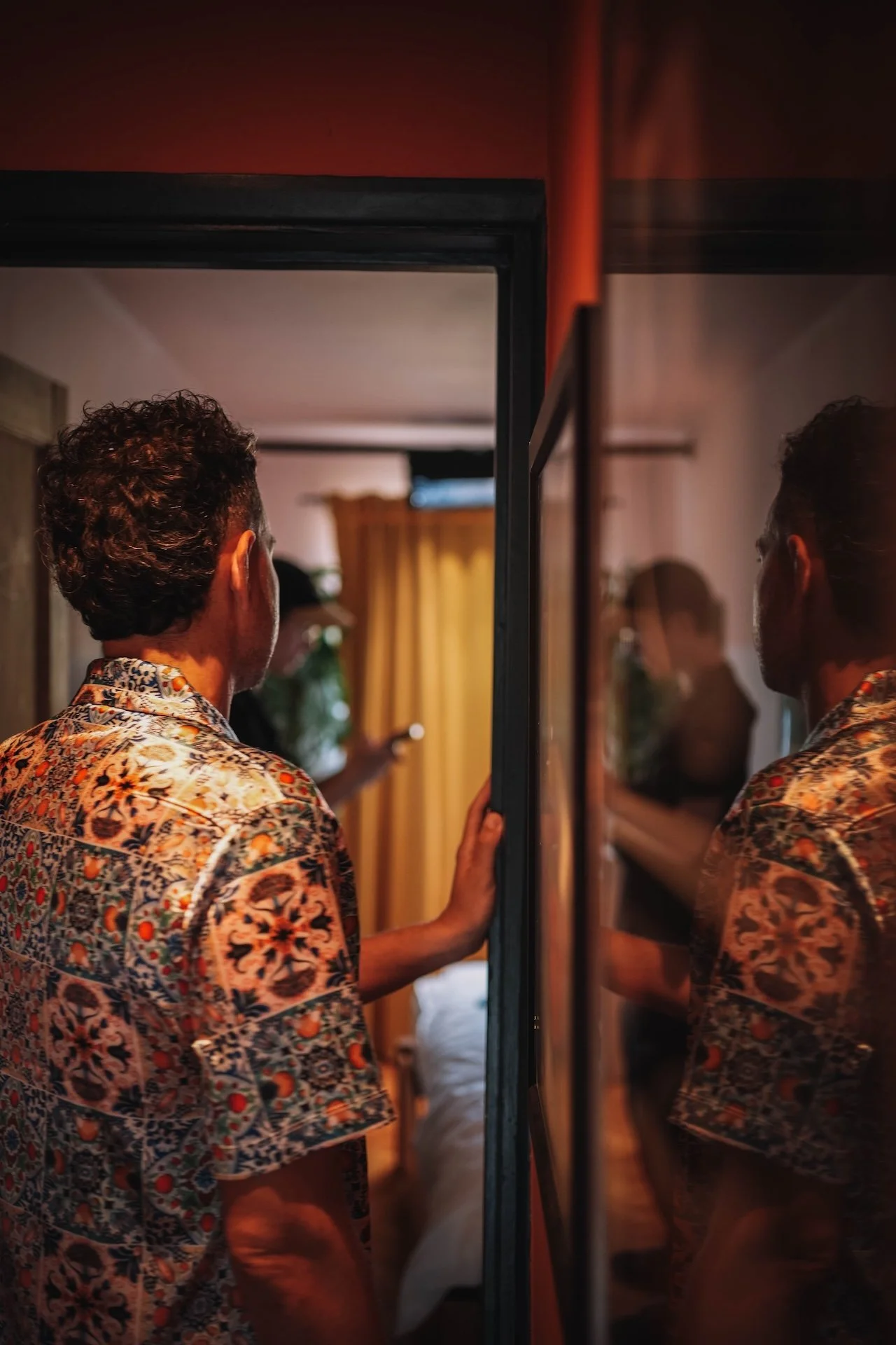 A person with curly hair wearing a colorful, patterned shirt looking at their reflection in a mirror in a dimly lit room.