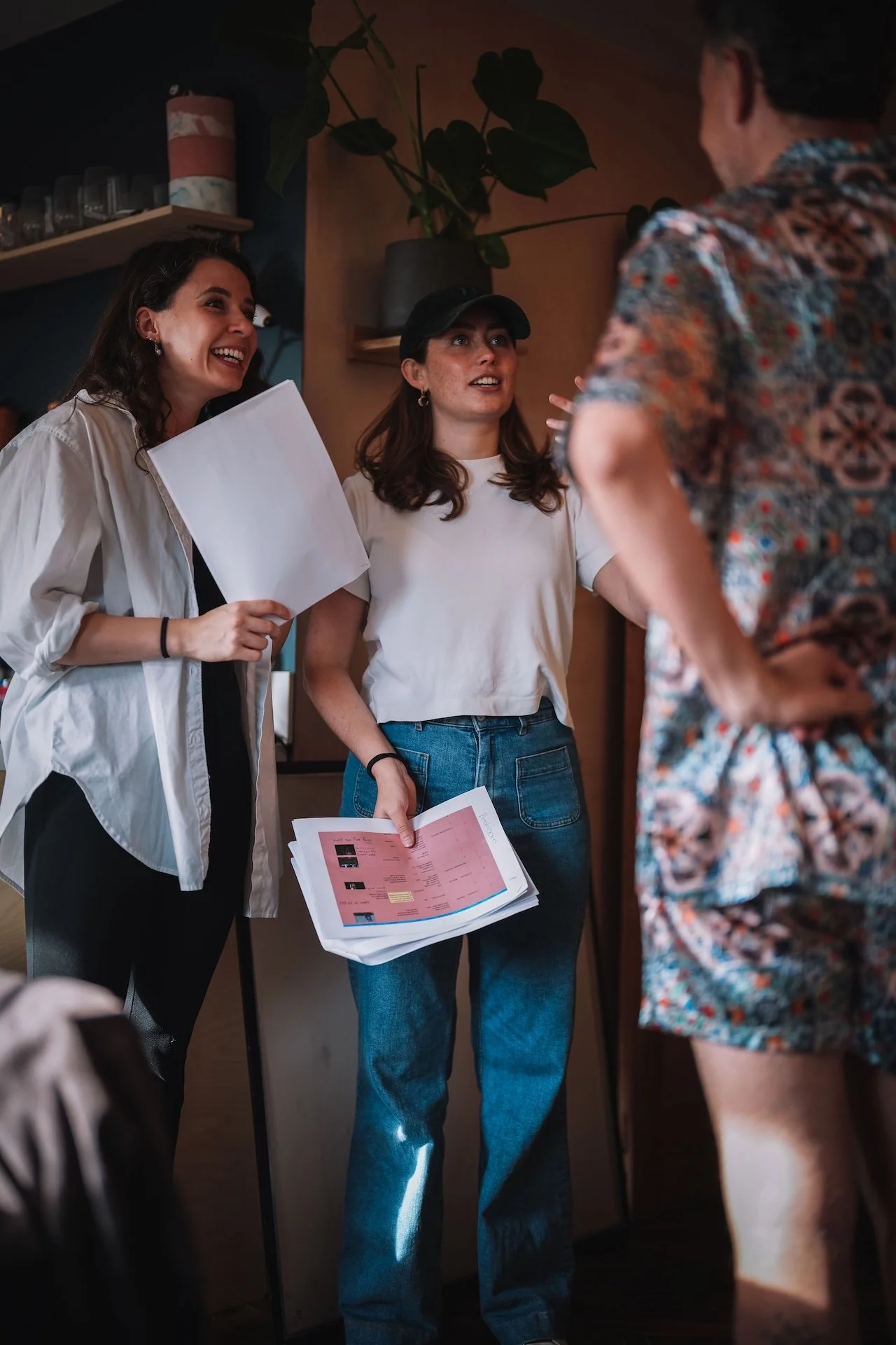 Three women in conversation in a cafe, one holding a pink printed sheet, two smiling and looking at the man in a patterned shirt.