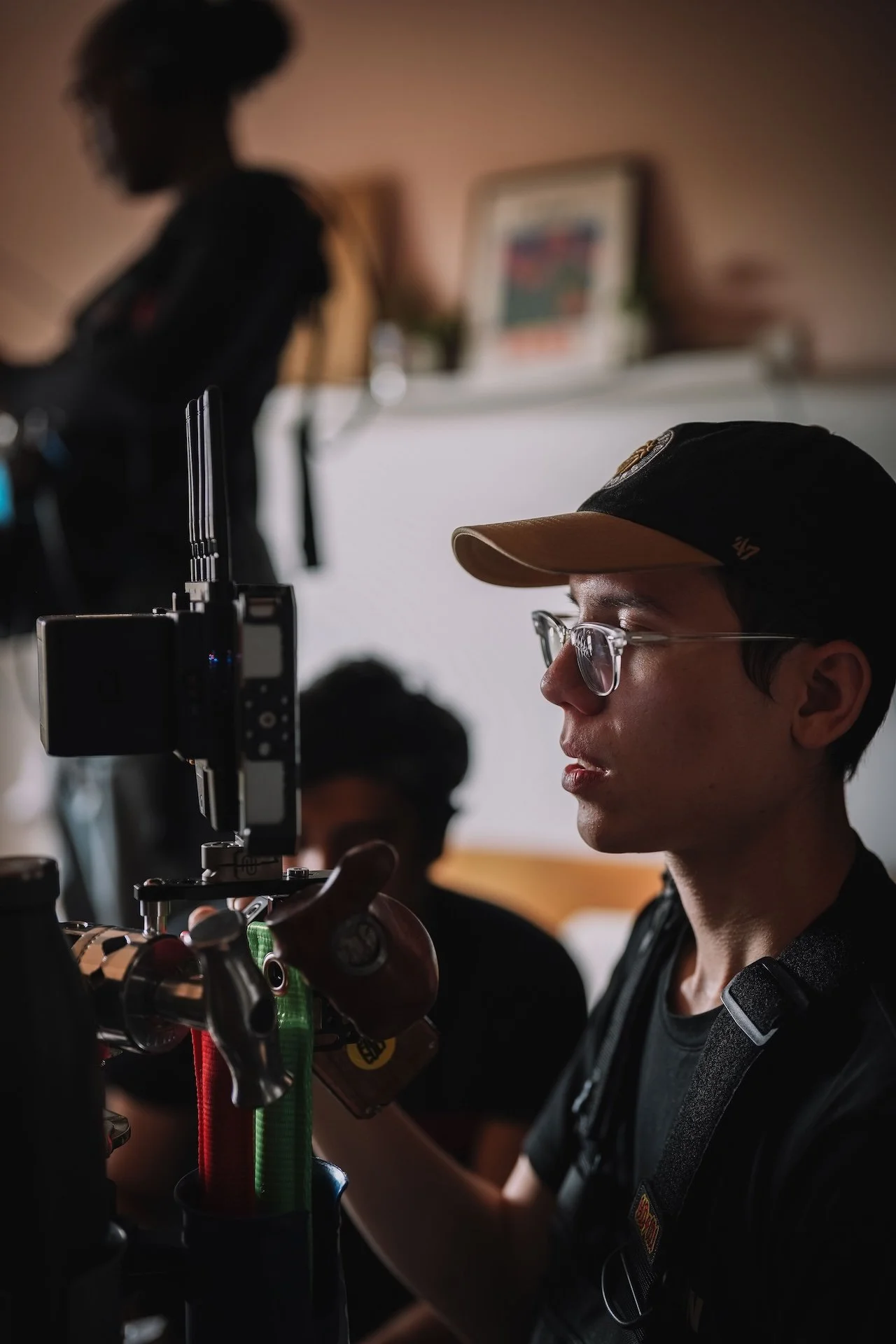 A person in glasses and a cap operating a microscope, with another person in the background, in a laboratory or workspace.