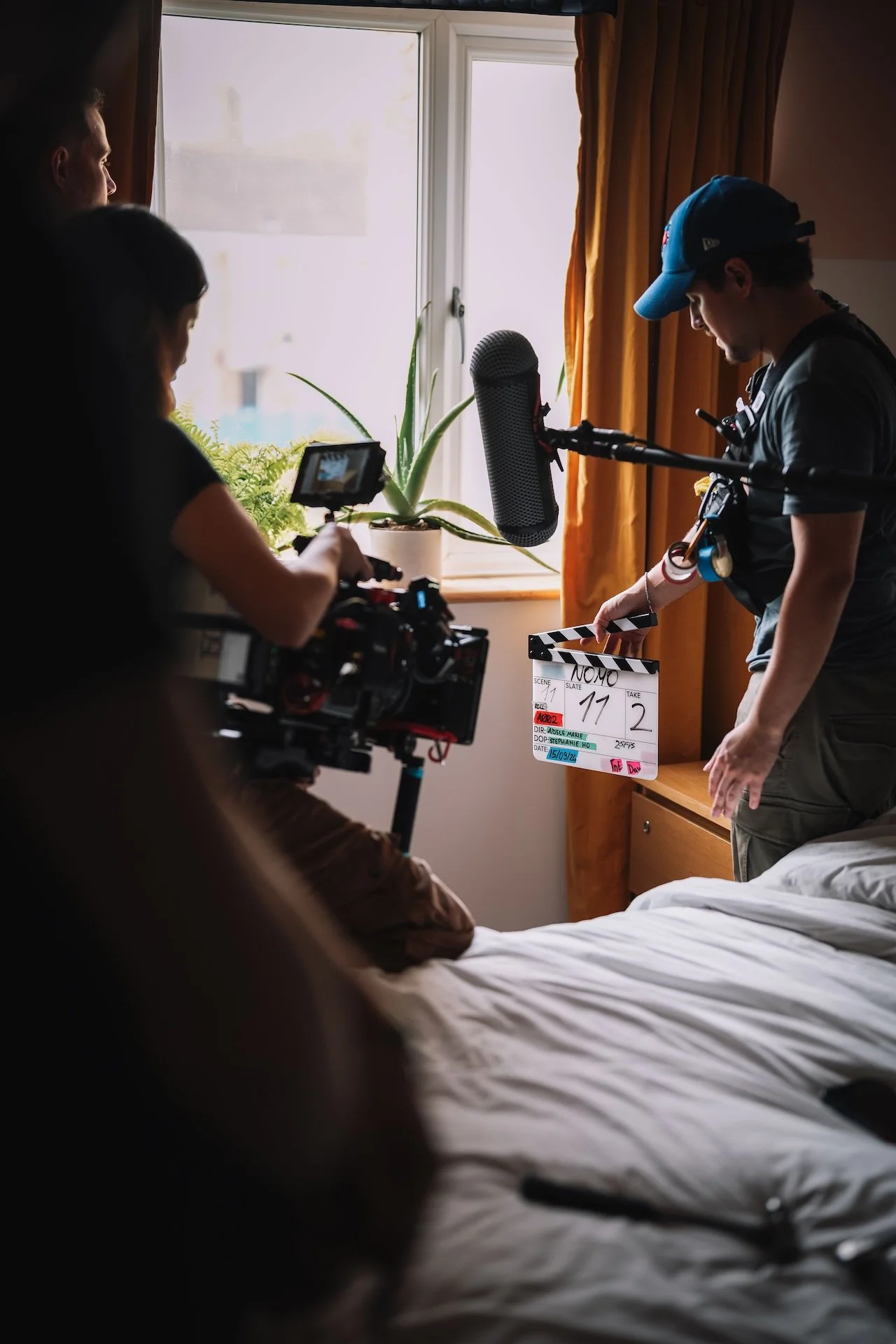 Scene from a film or TV show set, with a woman operating a camera and a man holding a clapperboard beside a person lying in bed near a window with curtains.