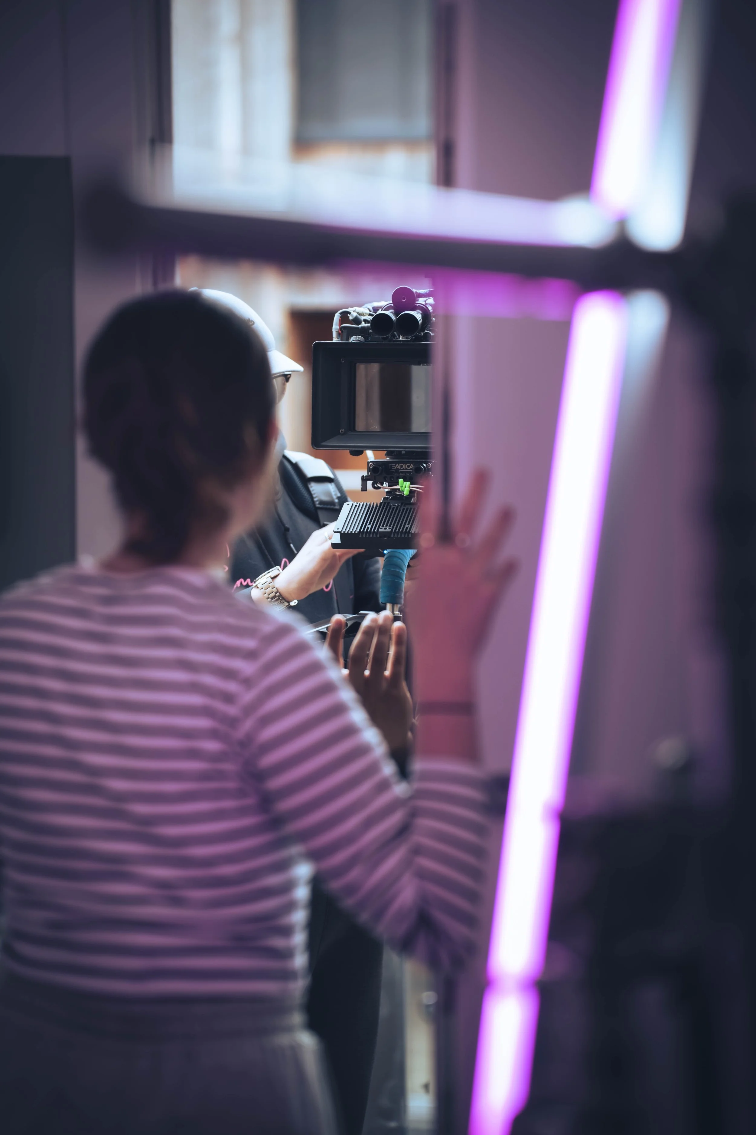 A person in a pink and white striped shirt stands in front of a professional video camera setup, with purple and pink neon lights reflecting on surrounding surfaces.