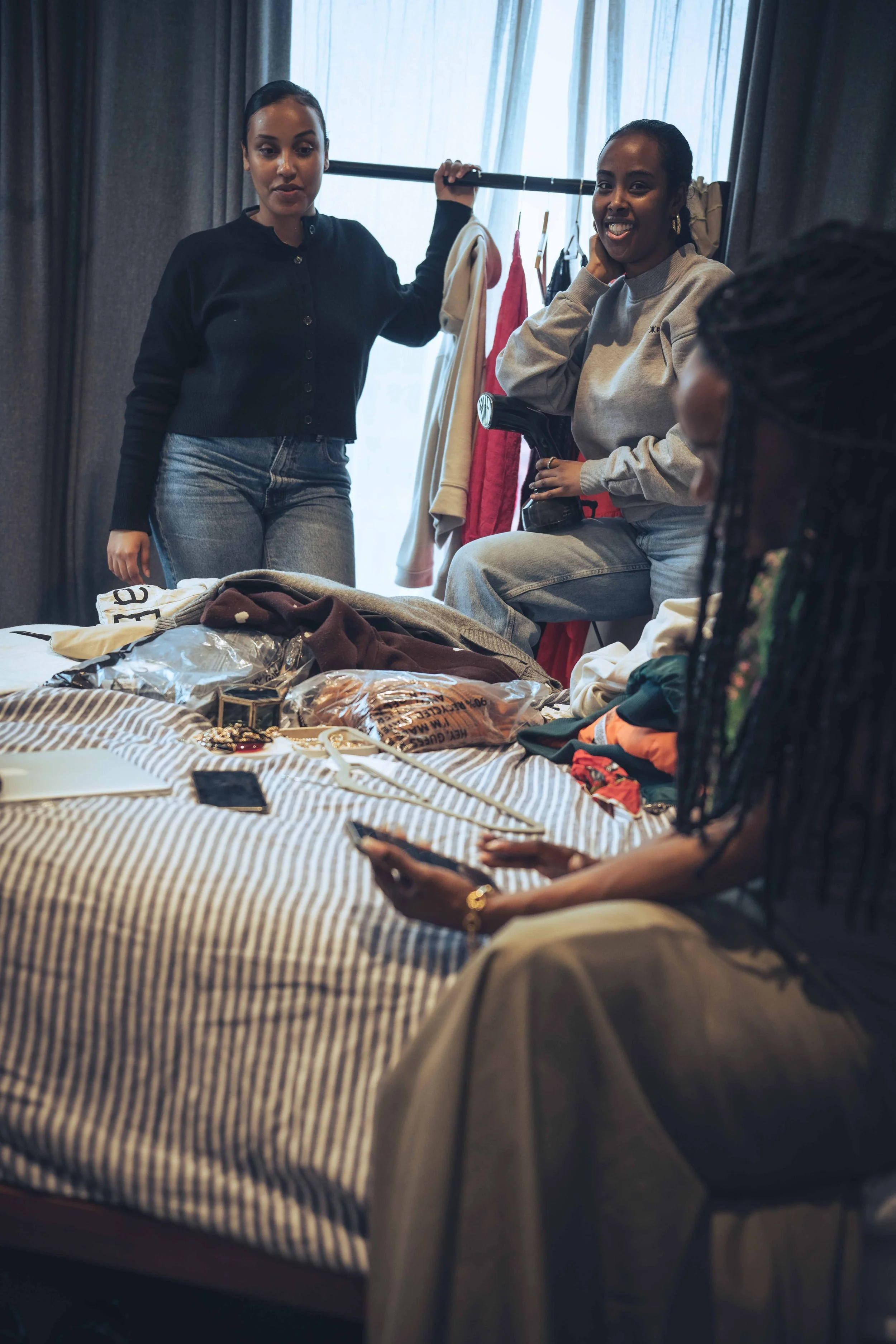 Women socializing in a room with clothes and accessories on a bed in the foreground. One woman is standing and another is sitting holding a camera, while a third woman is sitting and looking at her phone.