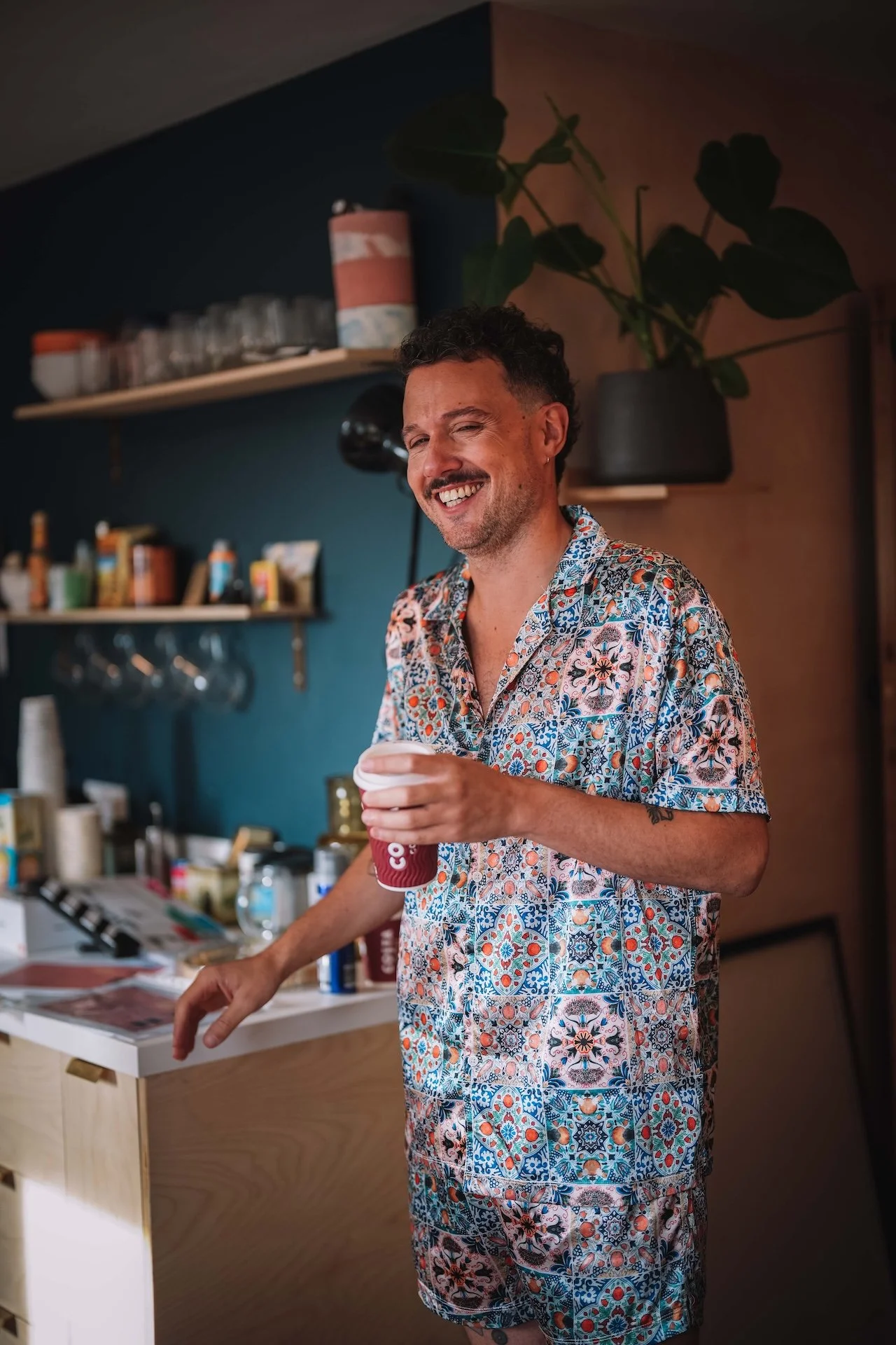 A man with short curly hair and a beard, wearing a colorful short-sleeve shirt and matching shorts, is smiling and holding a red cup while standing in a kitchen.