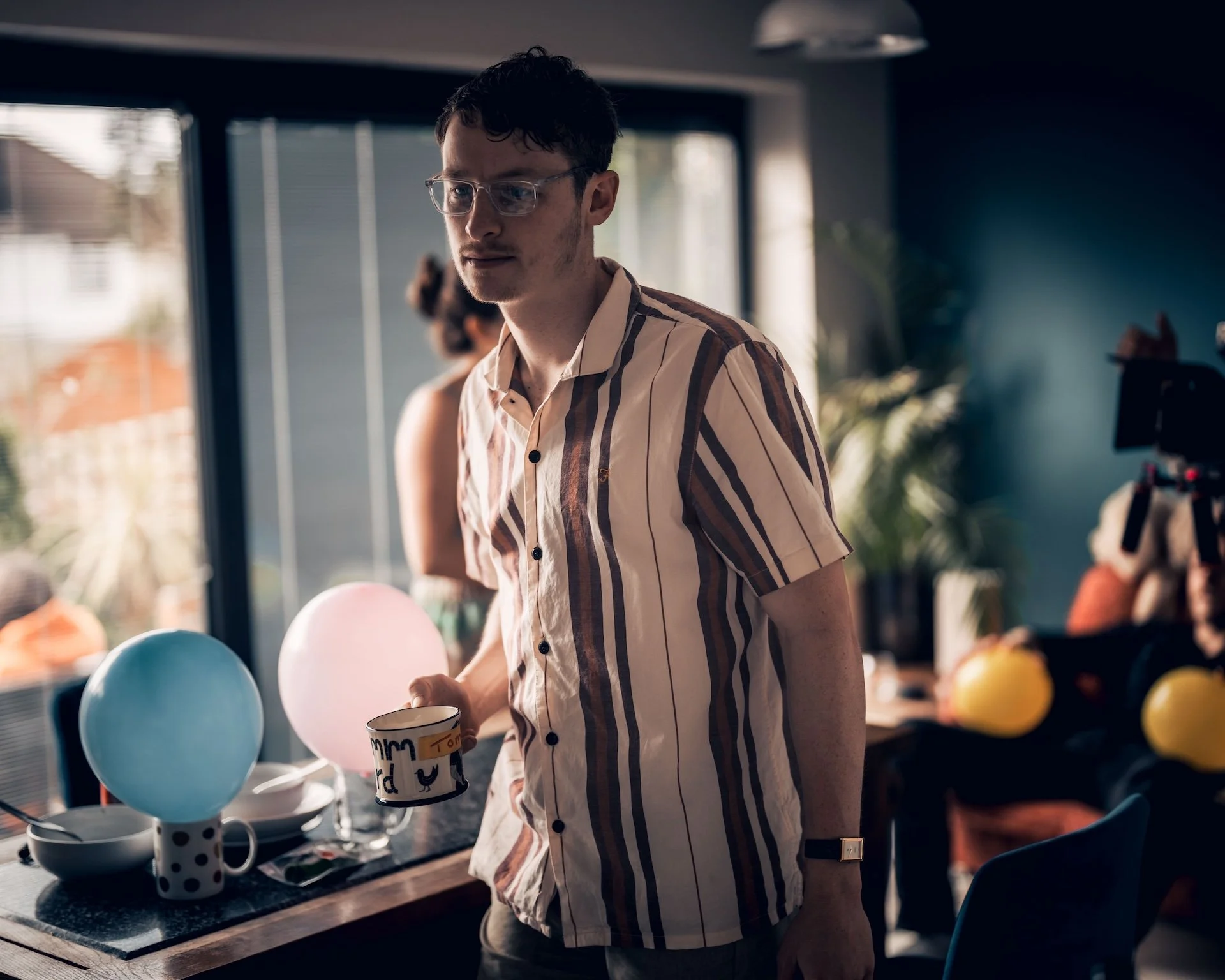 Young man with dark hair, glasses, and a striped beige and dark brown shirt standing in a room decorated with birthday balloons and mugs, holding a coffee mug, with a window and other people in the background.