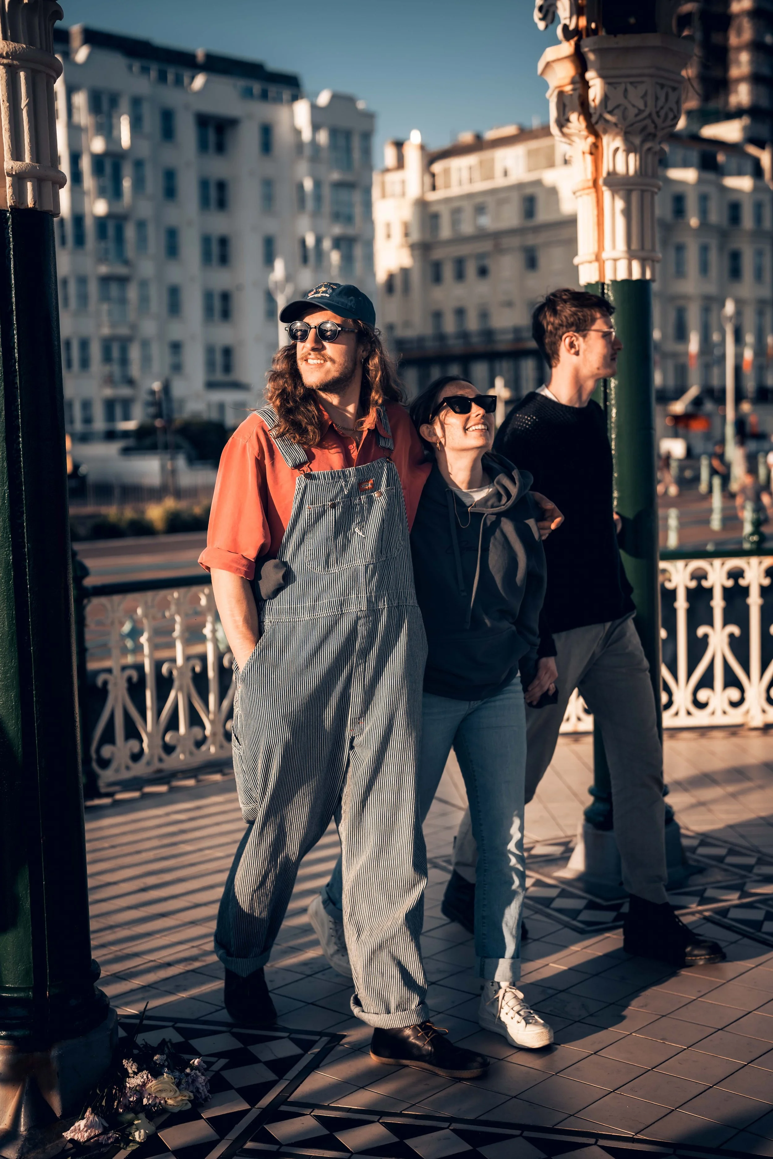 Three people standing on a patterned tile floor at an outdoor terrace or balcony, with ornate architectural details and buildings in the background. They are dressed casually and are looking in different directions, with warm sunlight illuminating th