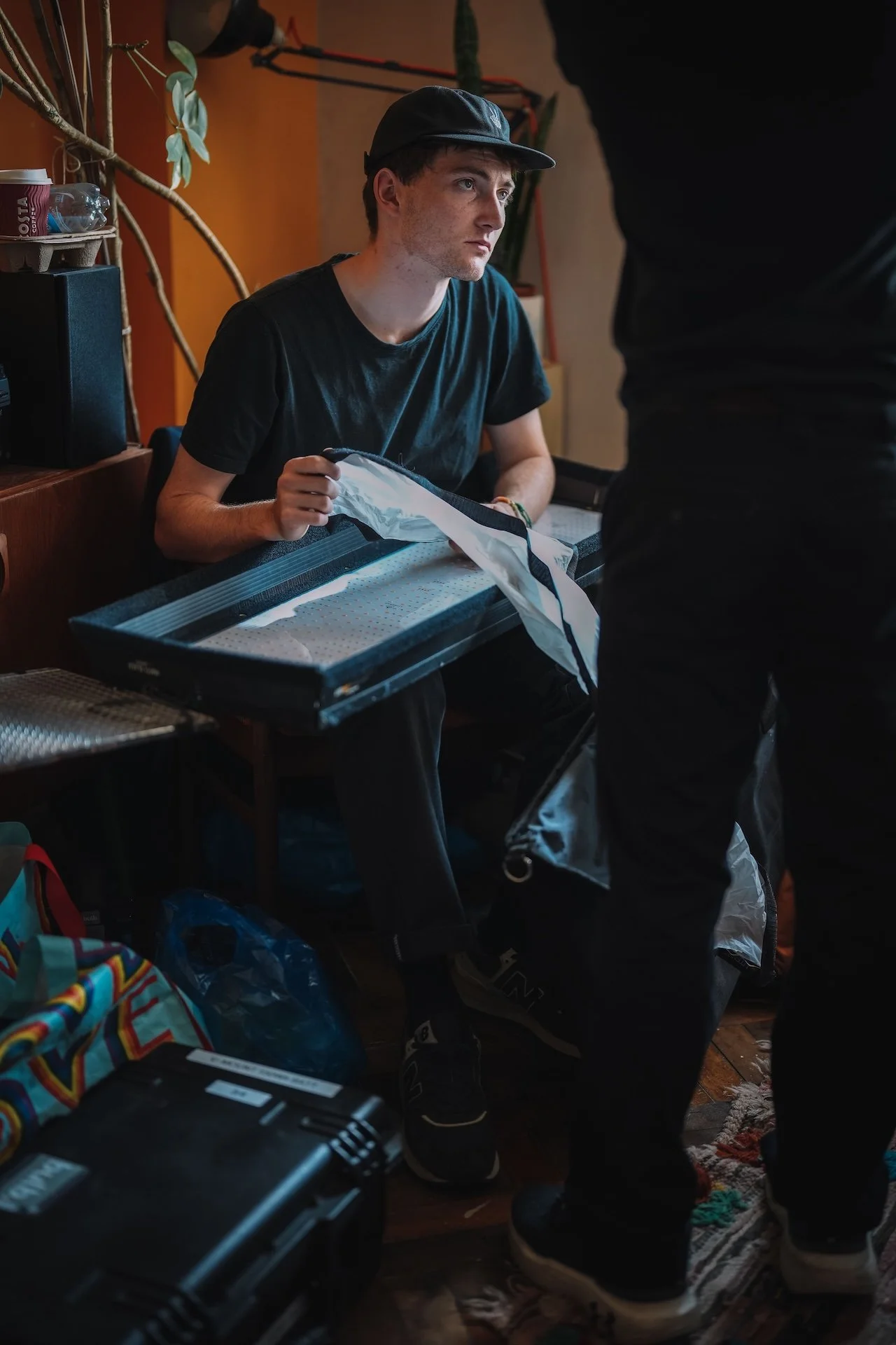 A young man sitting indoors at a table, looking to the side, wearing a black cap and black t-shirt, with various items, including a suitcase and bags, around him.