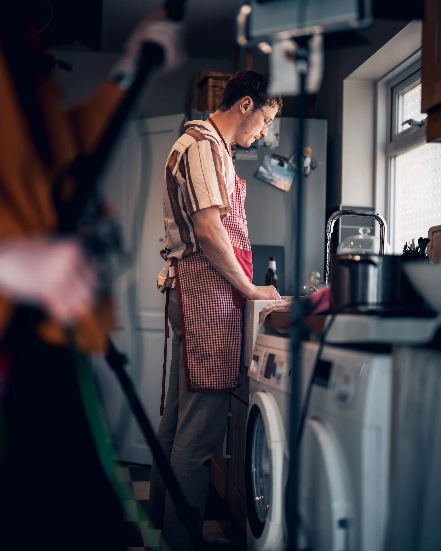 A man in an apron does laundry in a home laundry room near a window.