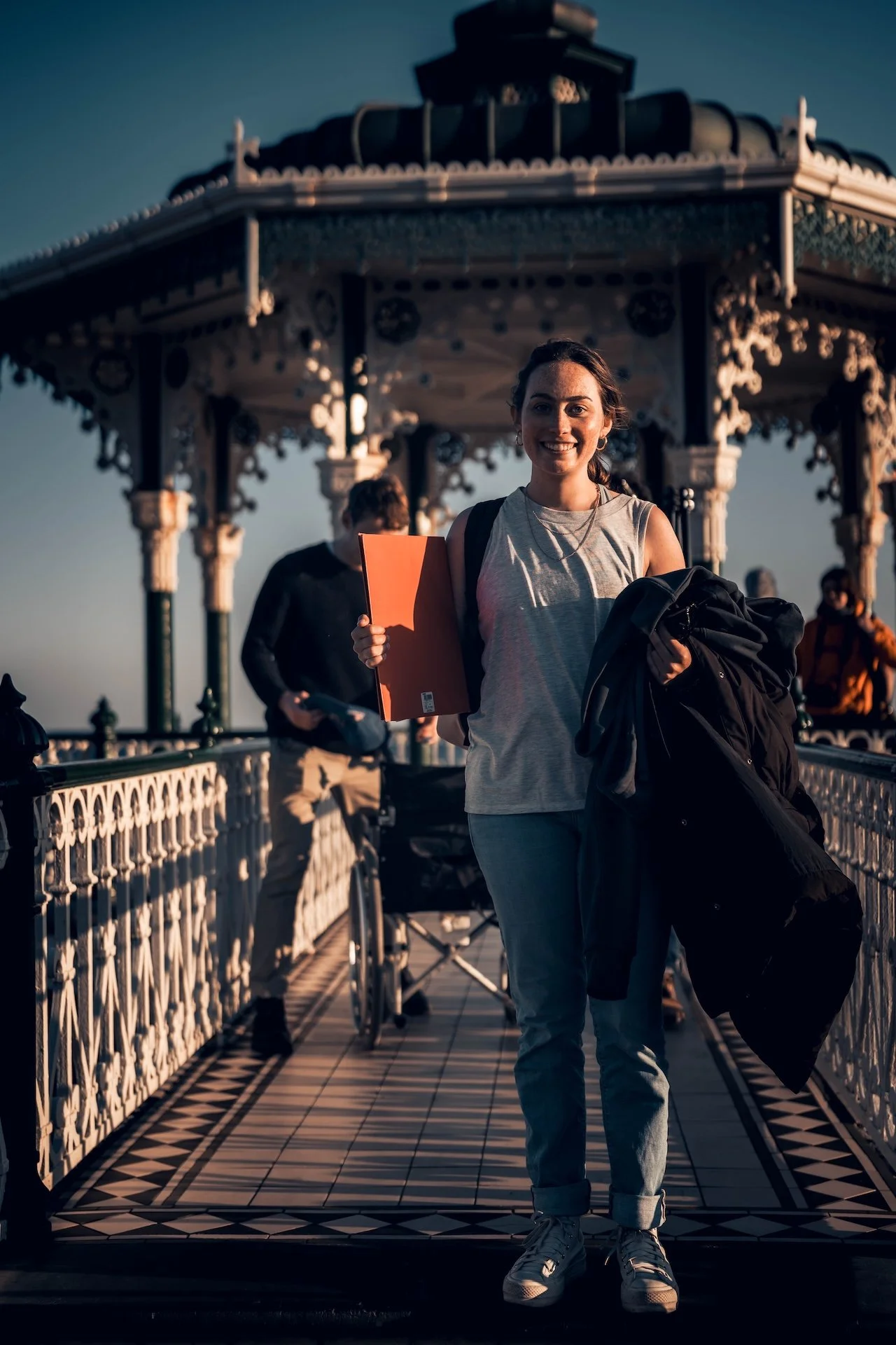 A woman smiling and holding a book and a jacket, walking on a decorated bridge with a pavilion in the background during sunset.