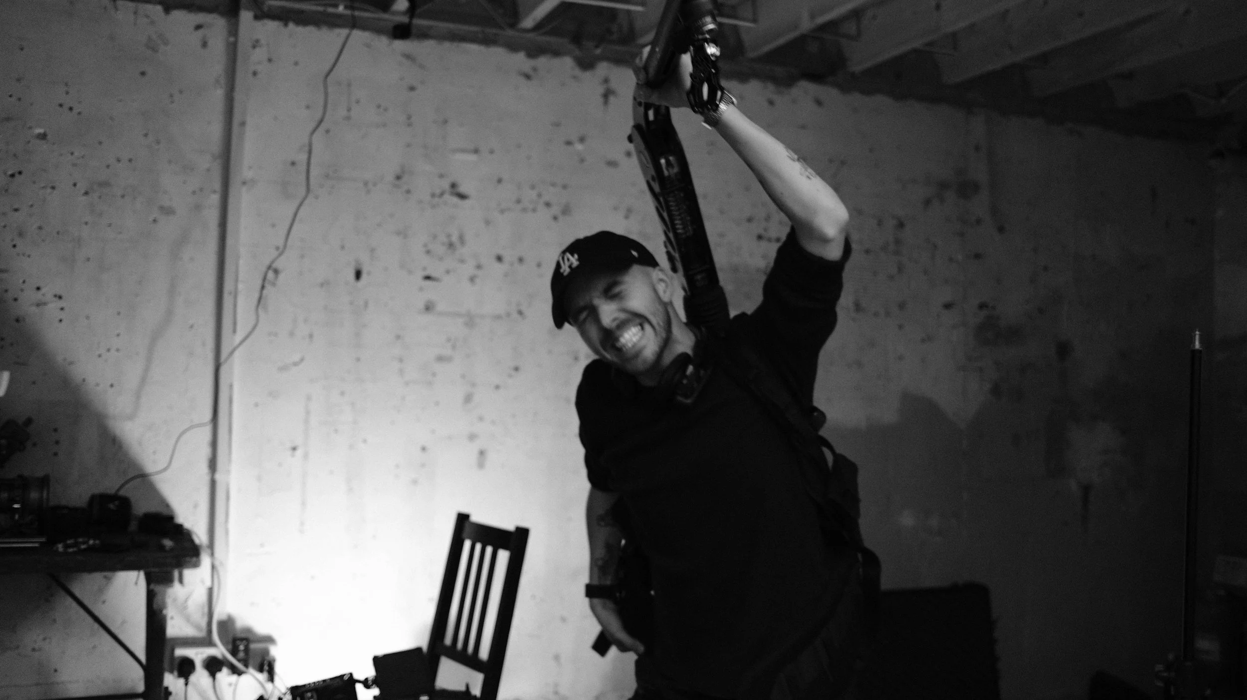 A man wearing a cap and black shirt, smiling widely, holding a camera rig above his head, standing in a room with unfinished walls and visible ceiling beams.