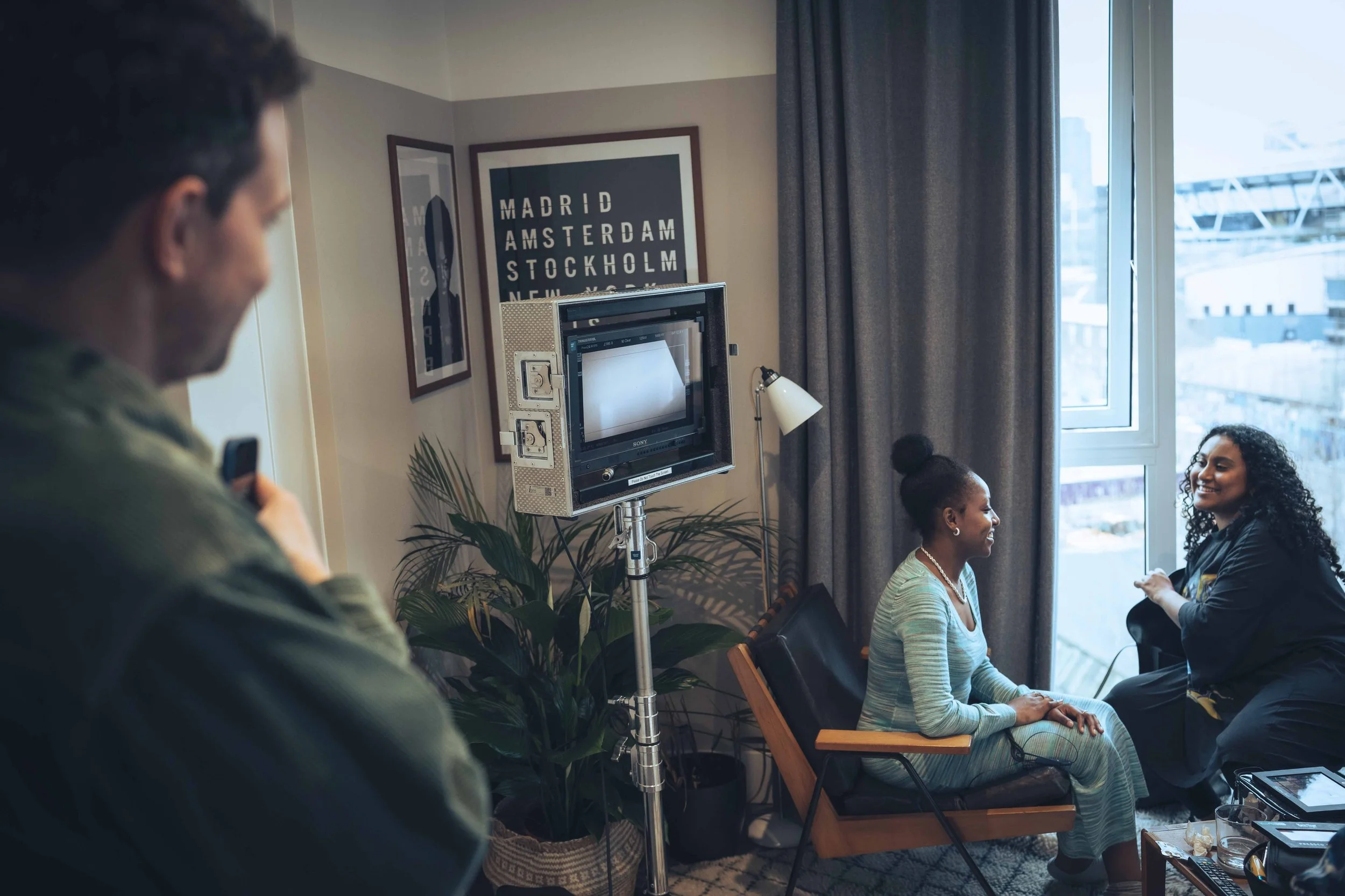 A group of women seated in a voting or interview setting, with two women smiling and talking to each other near a window. A man is in the foreground, slightly blurred, holding a device. A vintage television with a monitor and a studio light are also 
