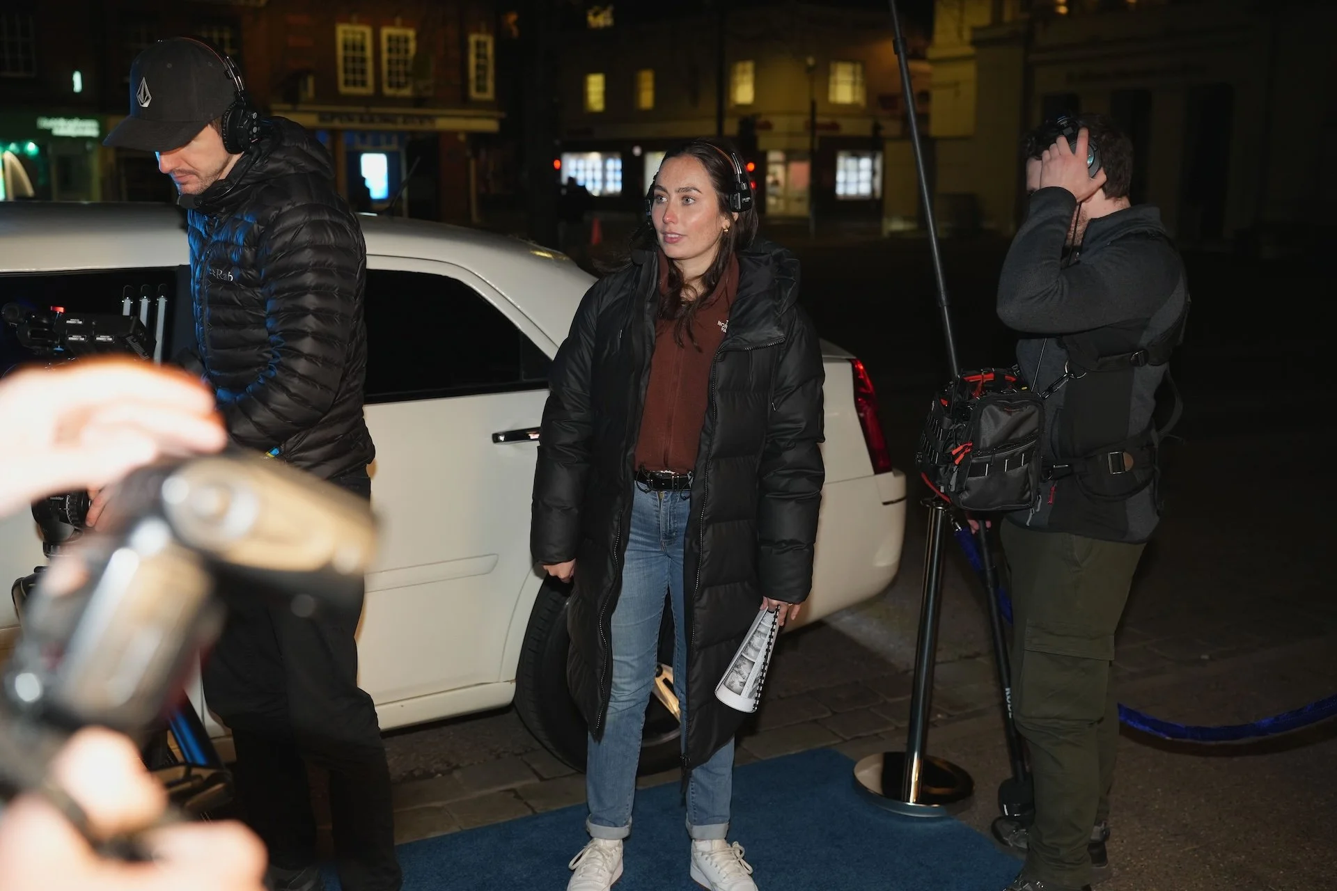 A woman standing next to a white vehicle, surrounded by a film crew at night on a city street.