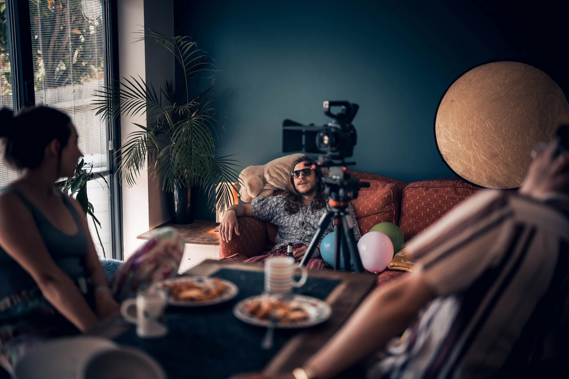 People celebrating a birthday indoors with balloons, cake, and a camera recording the event. One person with long hair and sunglasses is sitting on a couch, while others sit around a table.