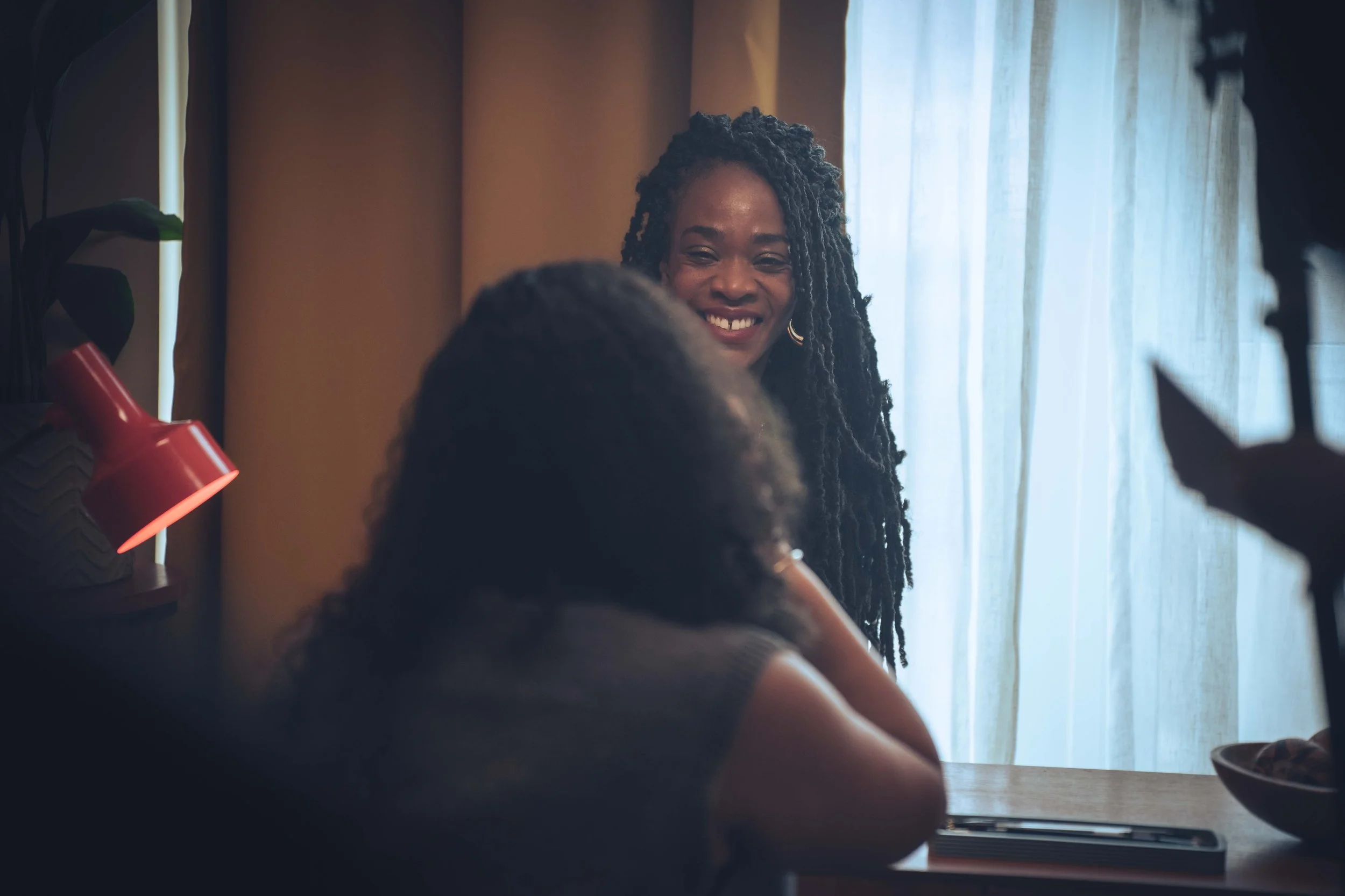 Smiling Black woman with dreadlocks, sitting by a window with sheer curtains, in a cozy room with warm lighting, possibly during daytime.