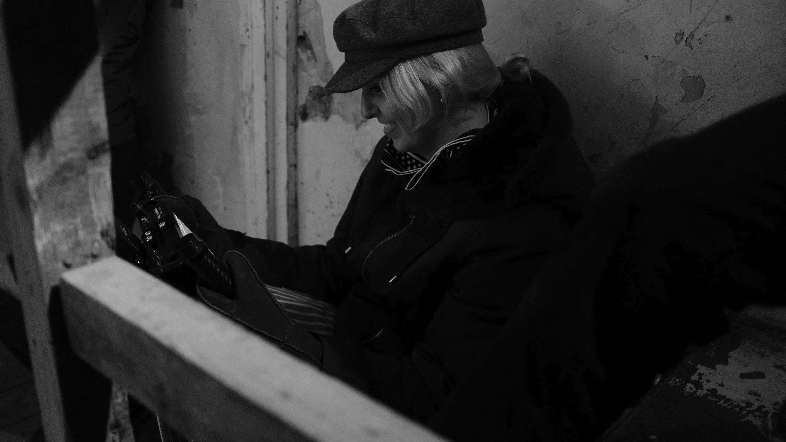 A woman wearing a hat and coat sitting on the floor and smiling while looking at a camera.