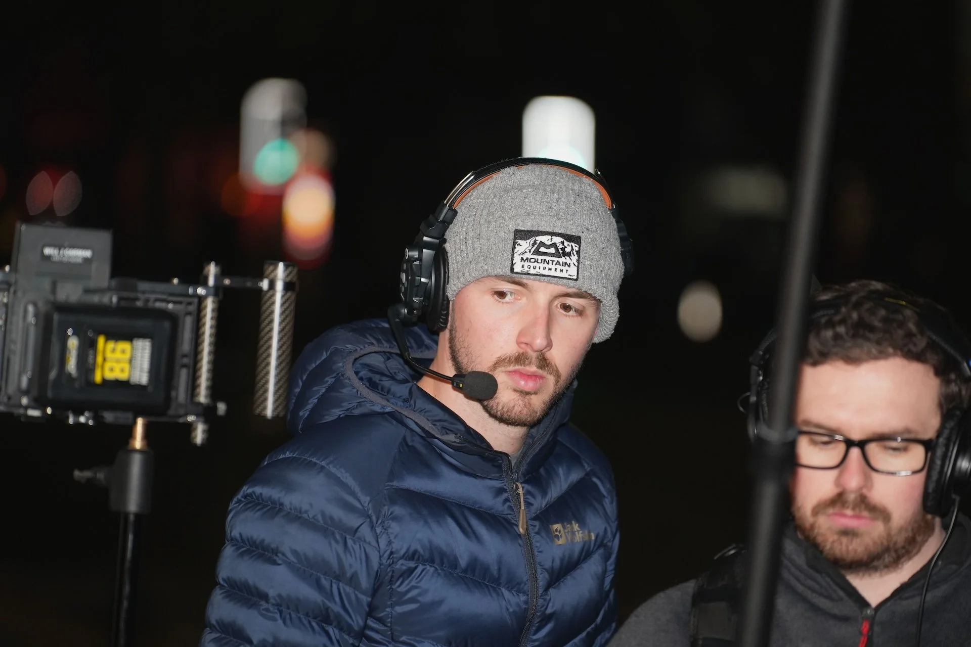 Two men with headphones and microphones, one with a gray beanie and blue jacket, and the other with glasses and a black jacket, working on a film or video production set at night.