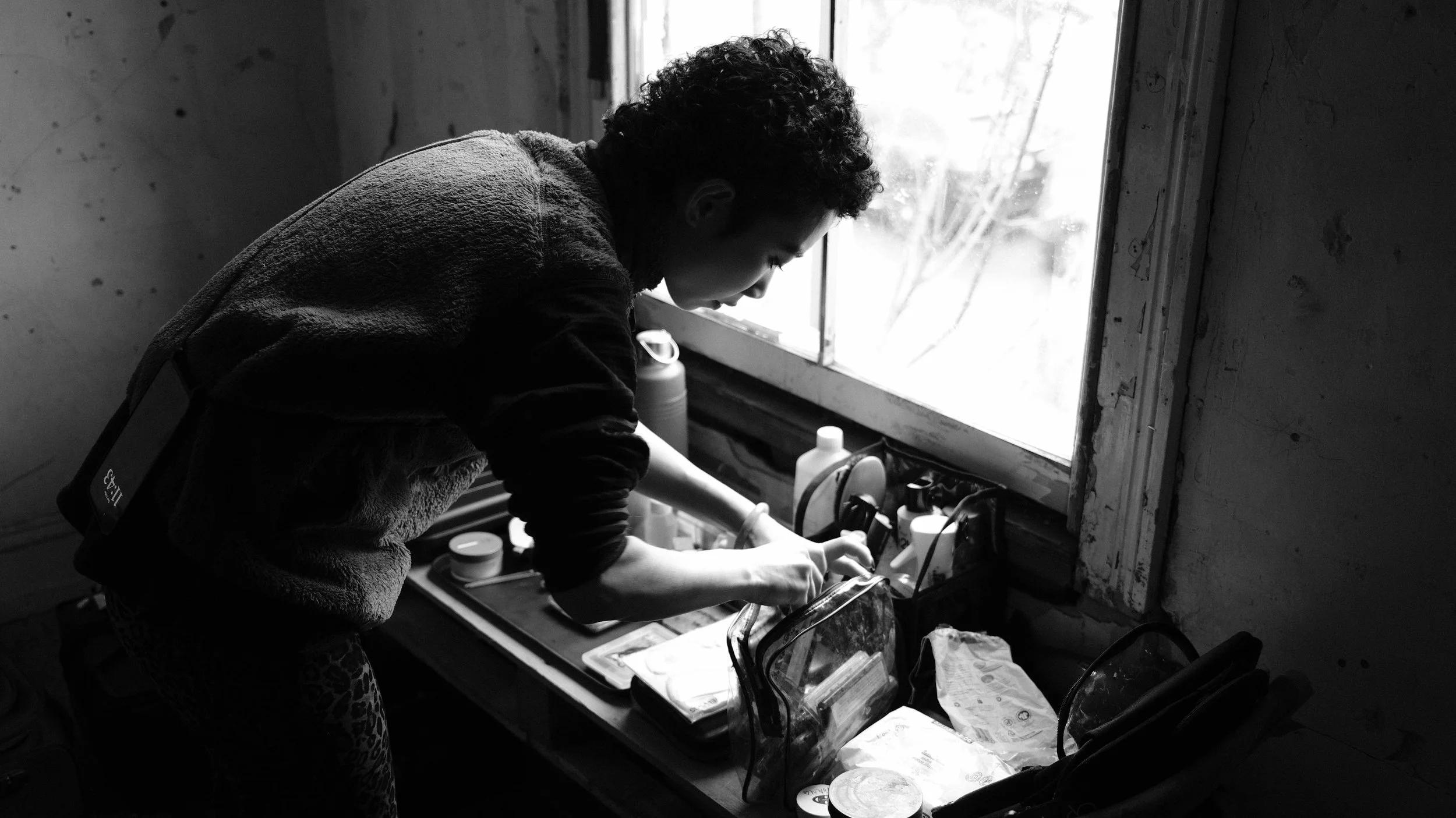 A person with short curly hair leaning over a sink or counter near a window, surrounded by bottles and containers, in a dimly lit room.