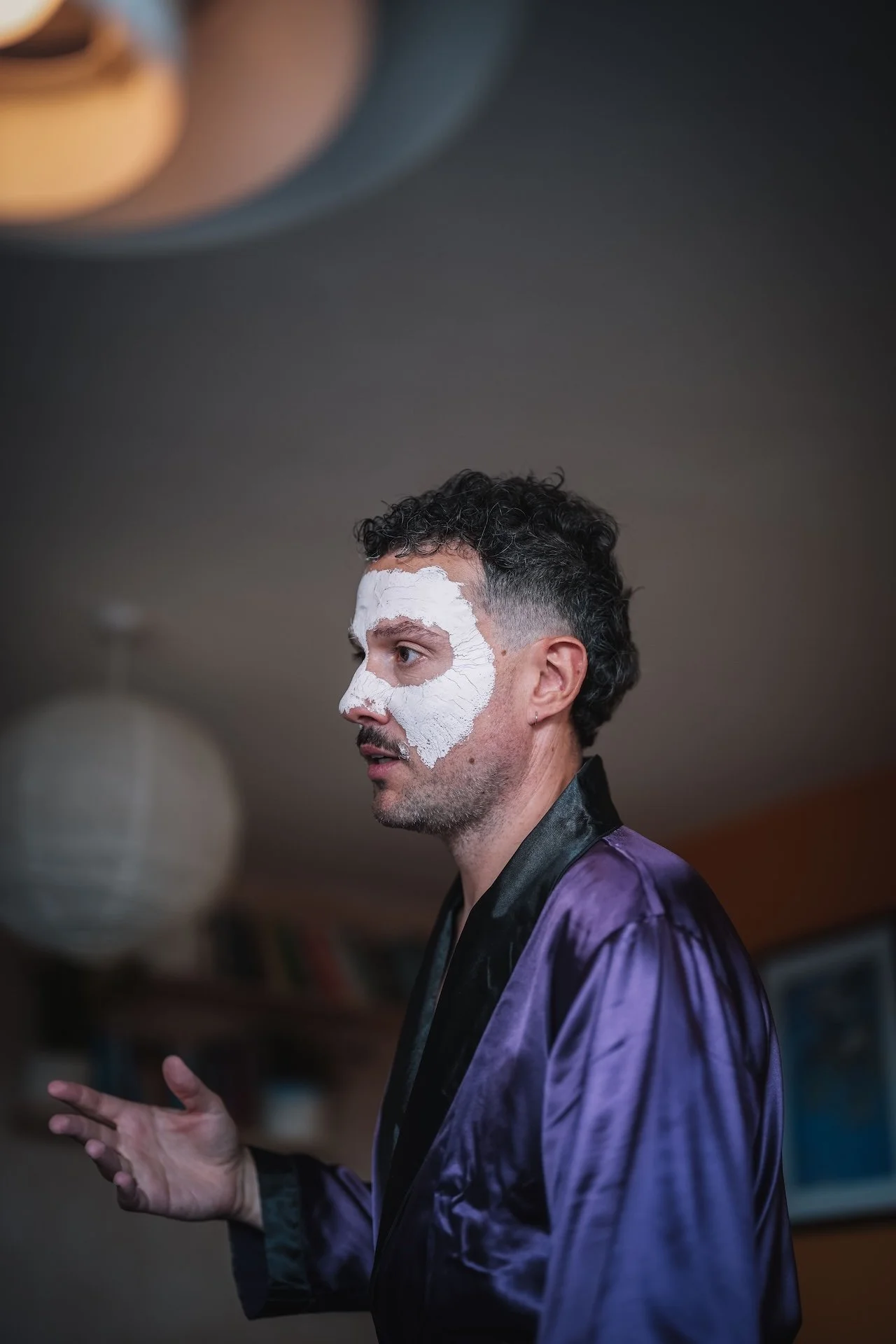 A man with curly dark hair applies a white facial mask while wearing a purple satin robe.