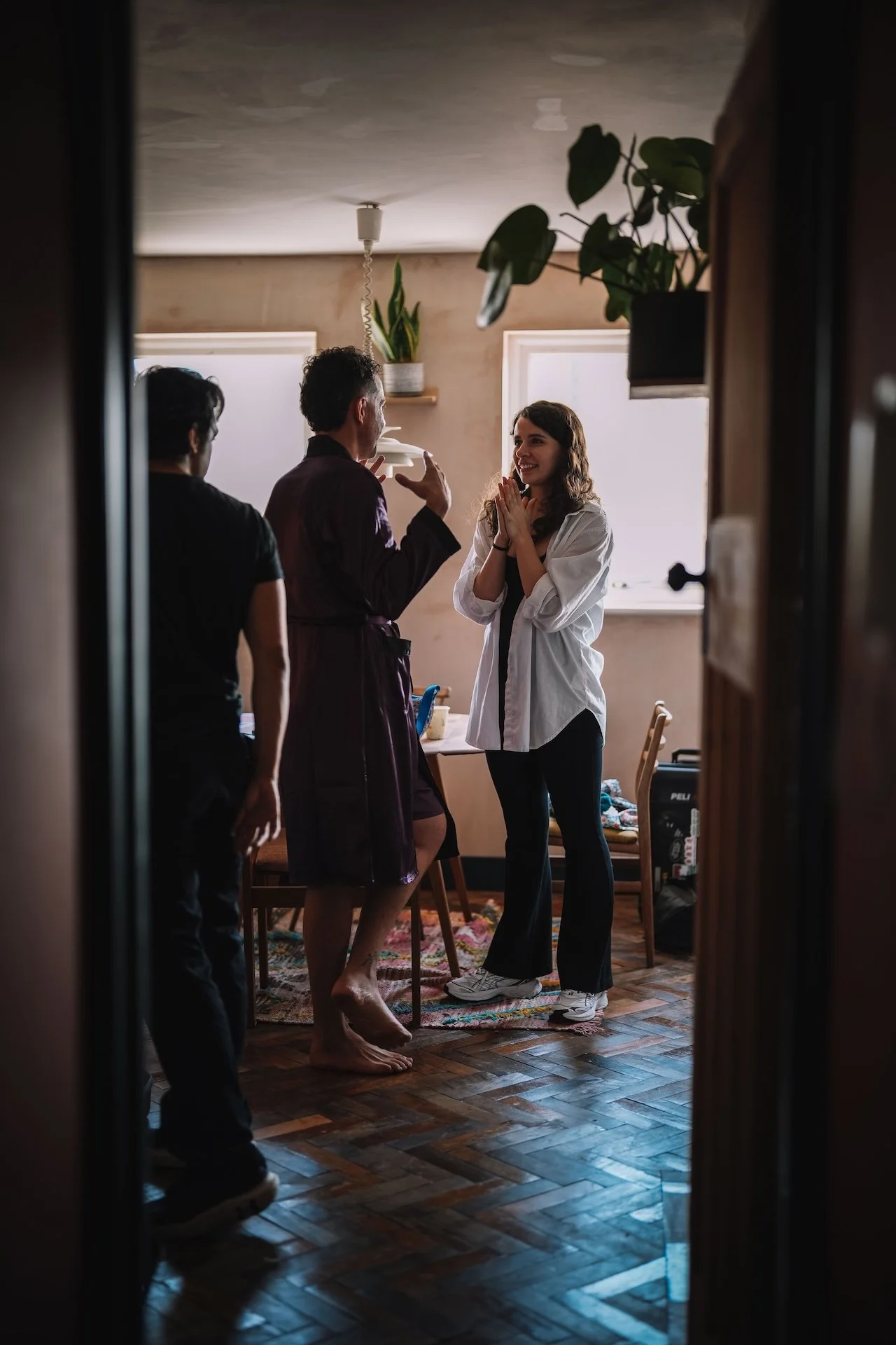 A young woman with long brown hair smiling and clapping hands, engaging with a man who has curly hair and a beard, in a cozy kitchen or dining area with natural light, plants, and wooden furniture.