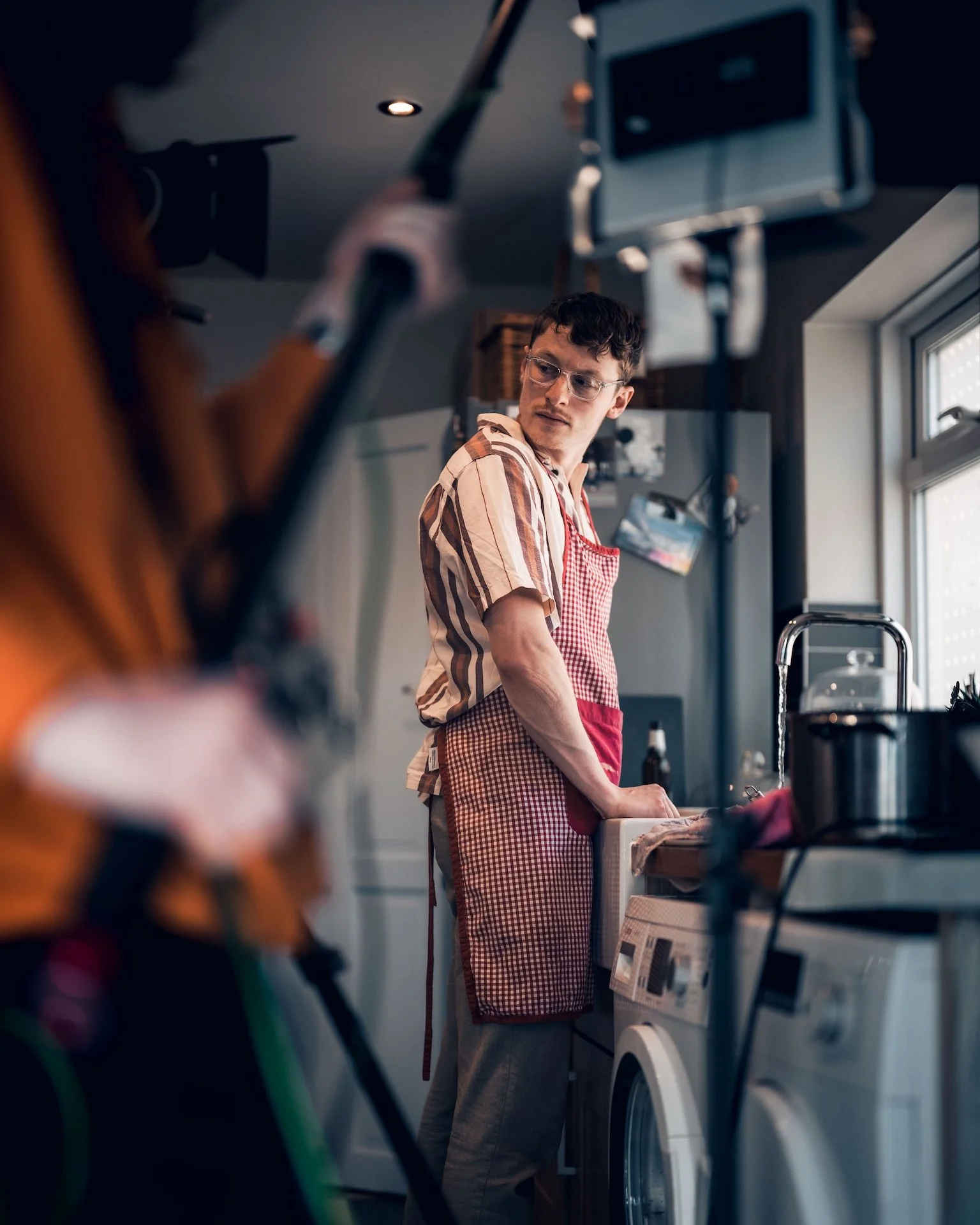 A young man with dark hair and glasses wearing a plaid shirt and red checkered apron doing laundry at a washing machine in a bright kitchen.