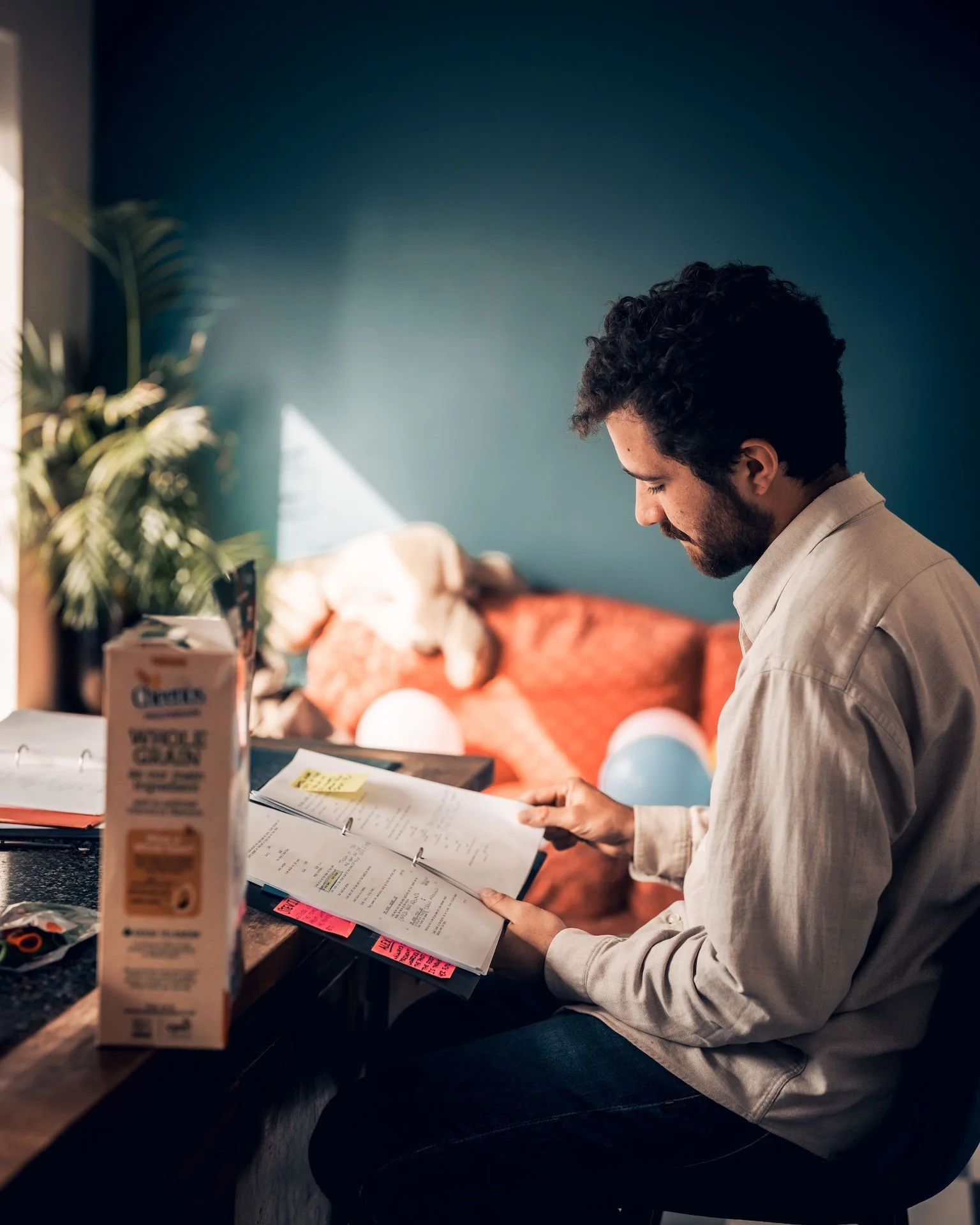 A man with dark curly hair and a beard is sitting at a cluttered counter, reading a binder with handwritten notes. In the background, a dog is resting on a patterned orange sofa in a cozy room with a teal wall. Items on the counter include a box of c