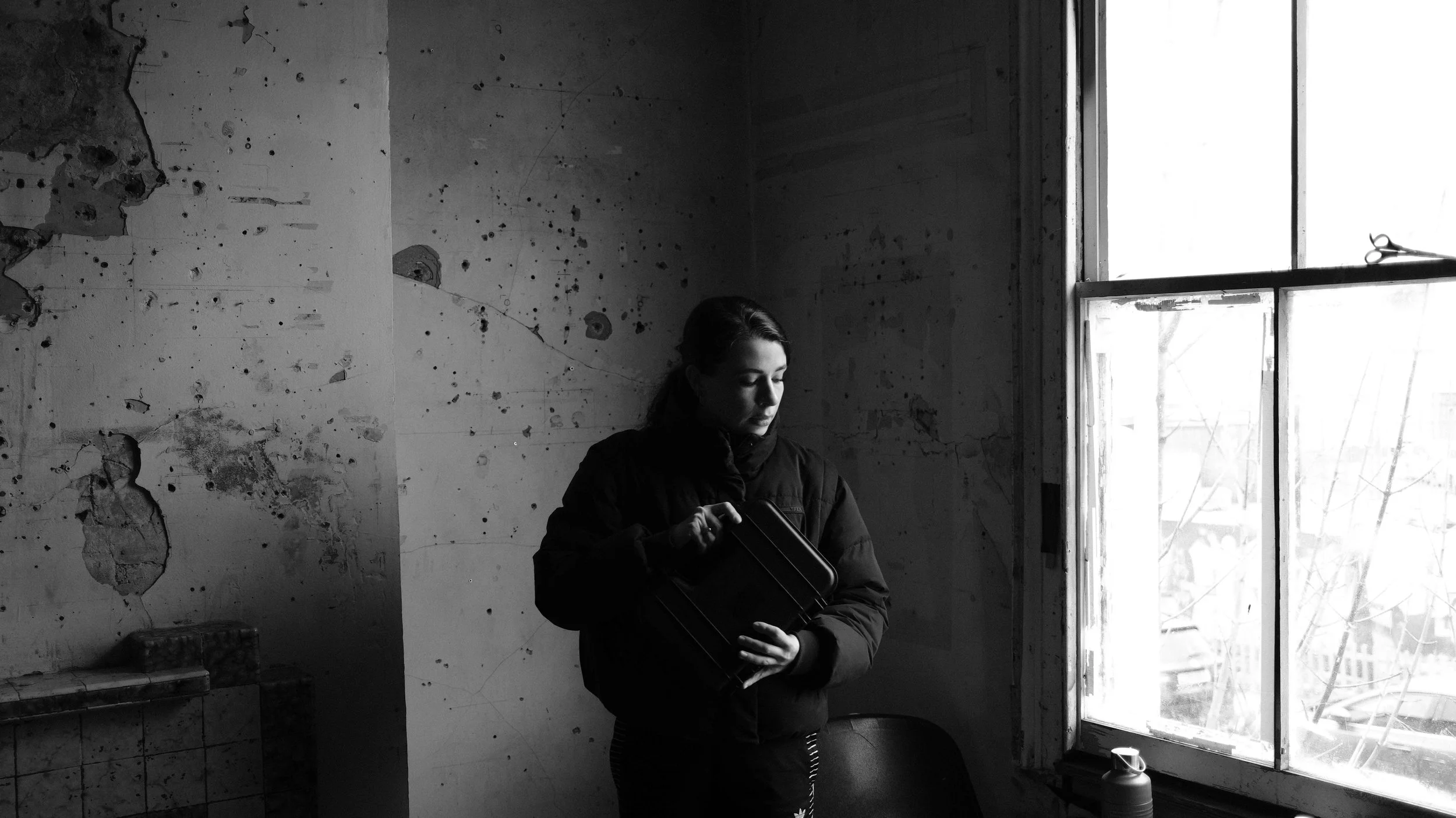 A woman in a dark jacket stands inside a room, holding a small suitcase and looking down. The room has a peeling wall with holes and a large window with light coming through, revealing some trees and a building outside.
