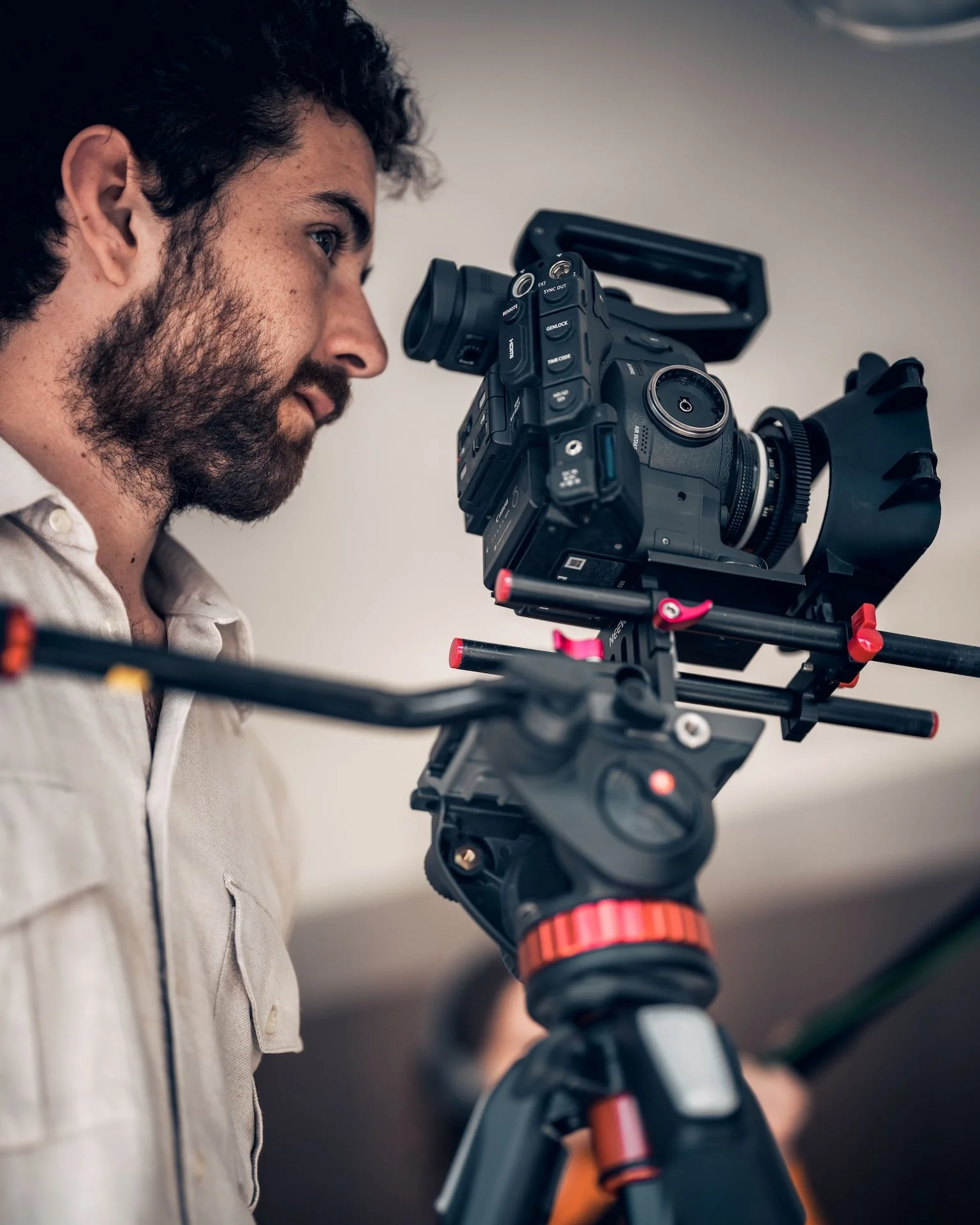 A man with dark, curly hair and a beard operating a professional video camera mounted on a tripod.