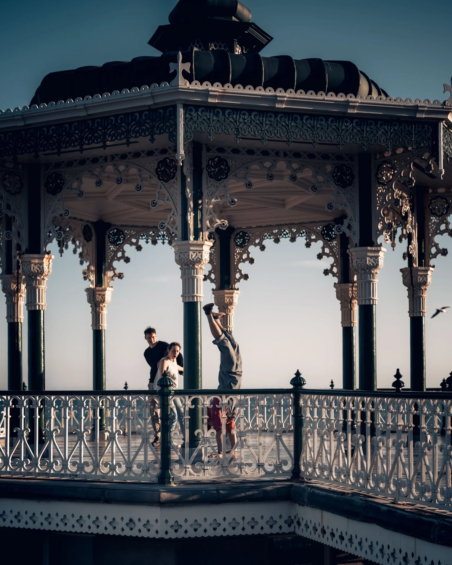 A decorative Victorian-style gazebo on a pier with ornate white railings and columns, featuring three people, two men and a woman, against a sky background.